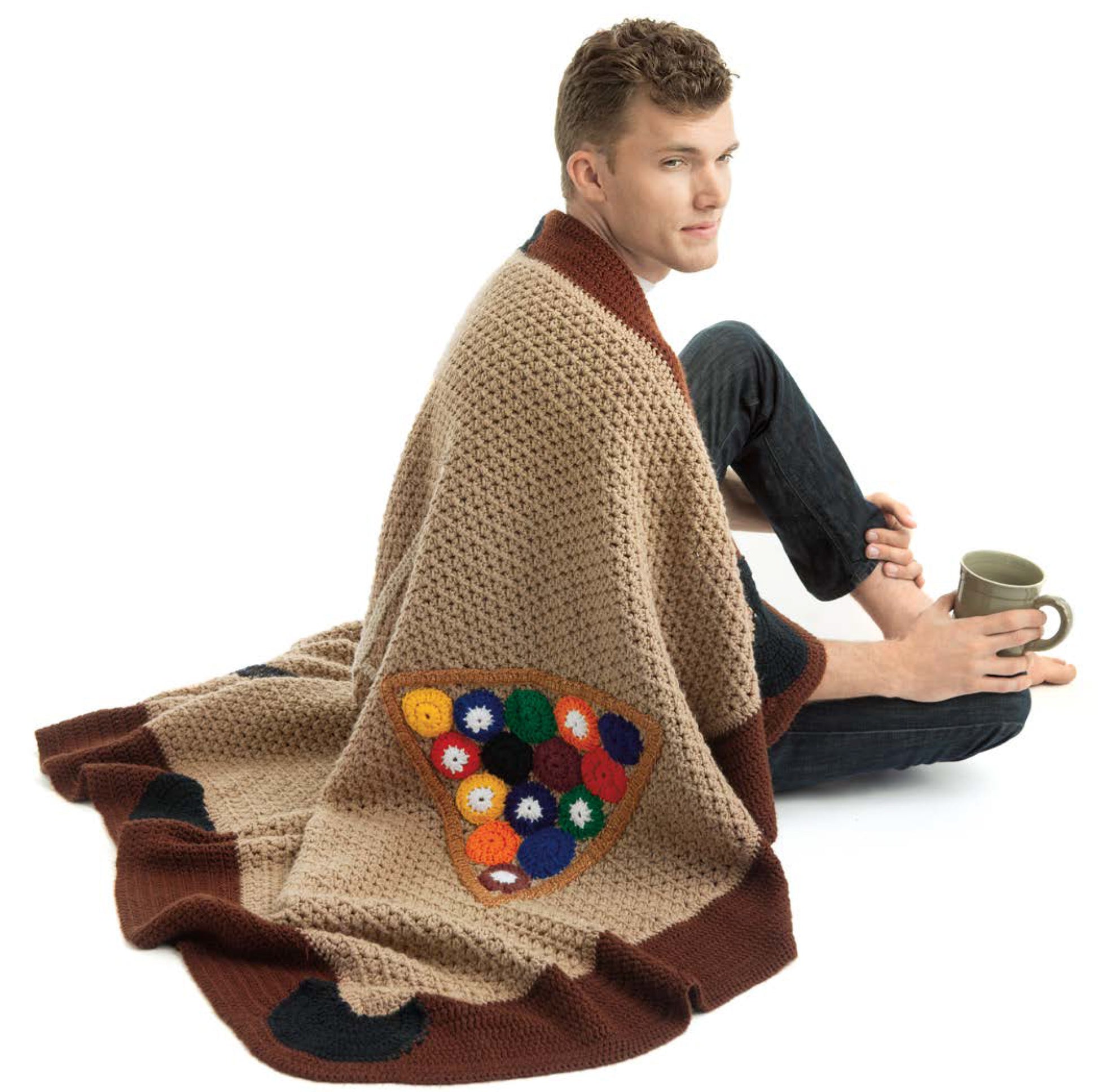 A man sits on the floor with a mug, wrapped in a beige crochet blanket featuring colorful accents—a perfect touch of unique style from the "Sportacular Afghans to Crochet Digital Download" by Leisure Arts for your man cave.