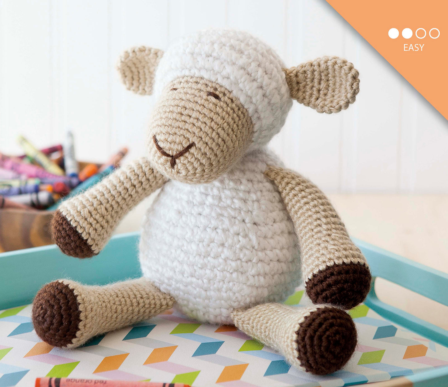 The Baby's Buddy Amigurumi by Leisure Arts, an easy crochet project ideal for beginners or fans of crochet baby toys, rests on a vibrant geometric surface with crayons in a tray nearby. An "easy" difficulty label appears at the top right.