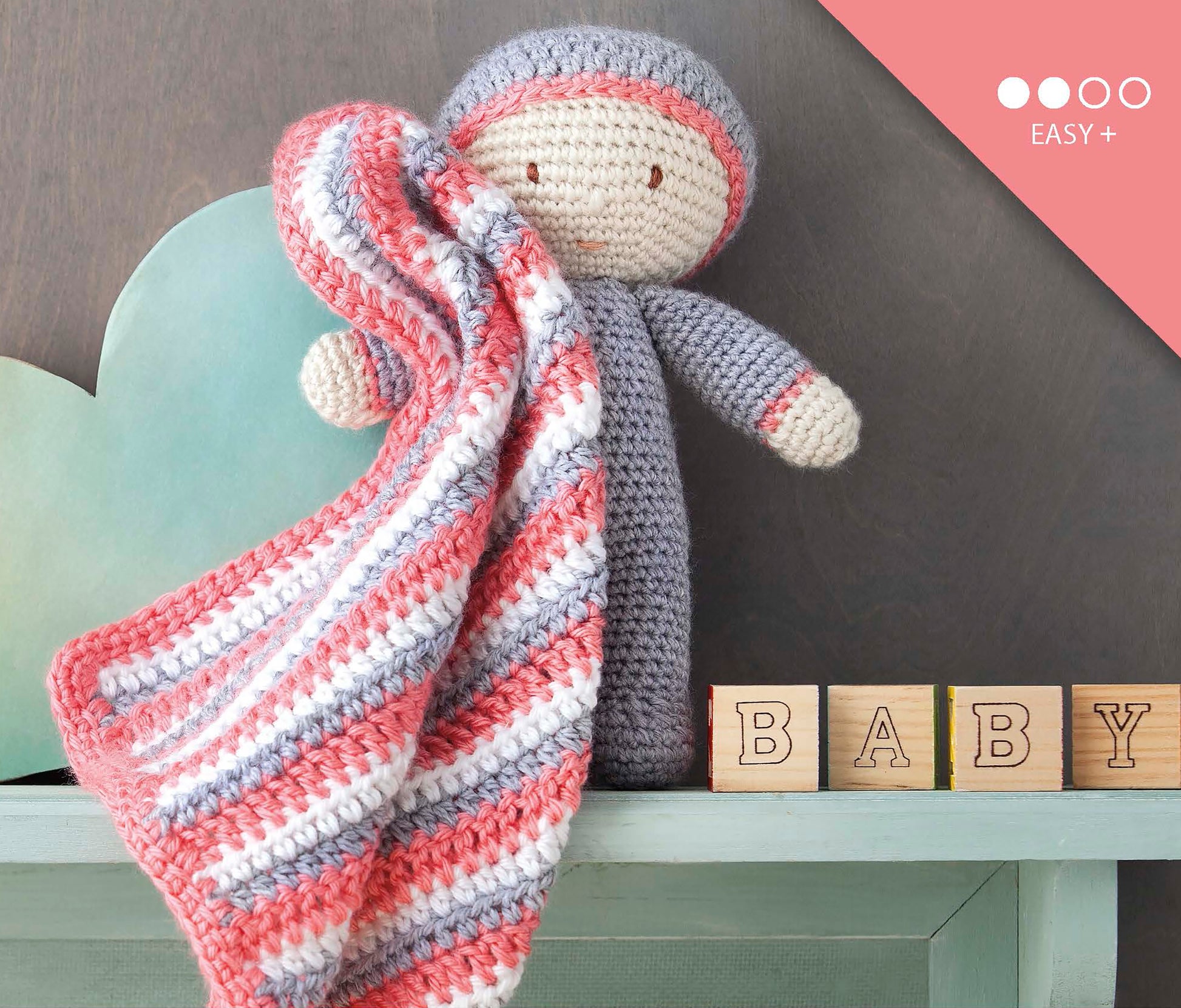 Baby's Buddy Amigurumi by Leisure Arts features a crocheted doll in gray holding a striped pink, white, and gray blanket, with pastel BABY blocks and a mint-green heart—ideal for crochet lovers or those seeking beginner patterns. Pink EASY+ label included.