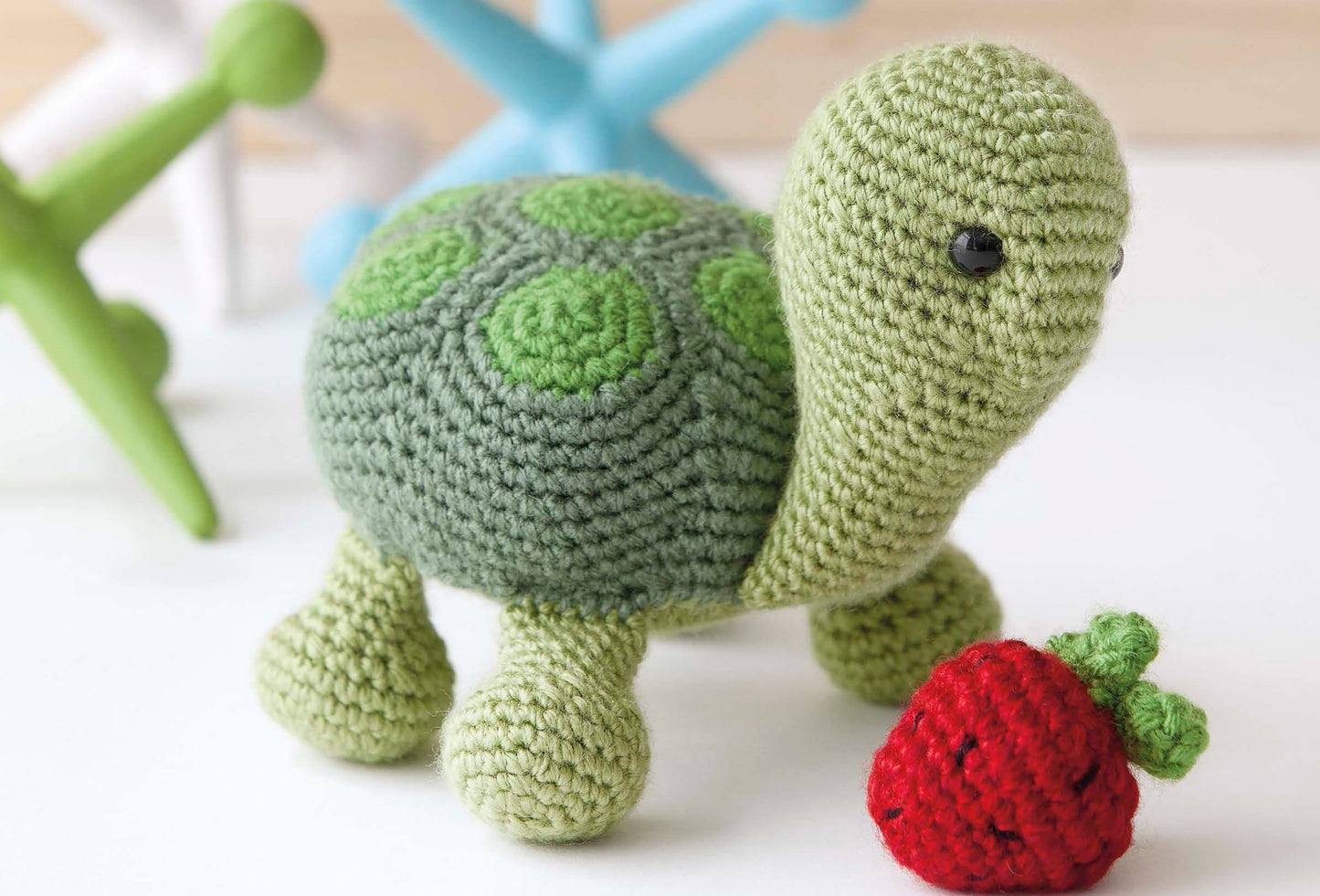 Baby's Buddy Amigurumi by Leisure Arts features a green crocheted turtle with a patterned shell and a red strawberry, perfect for crochet stuffed animal fans, shown on a white surface with colorful jacks blurred in the background.