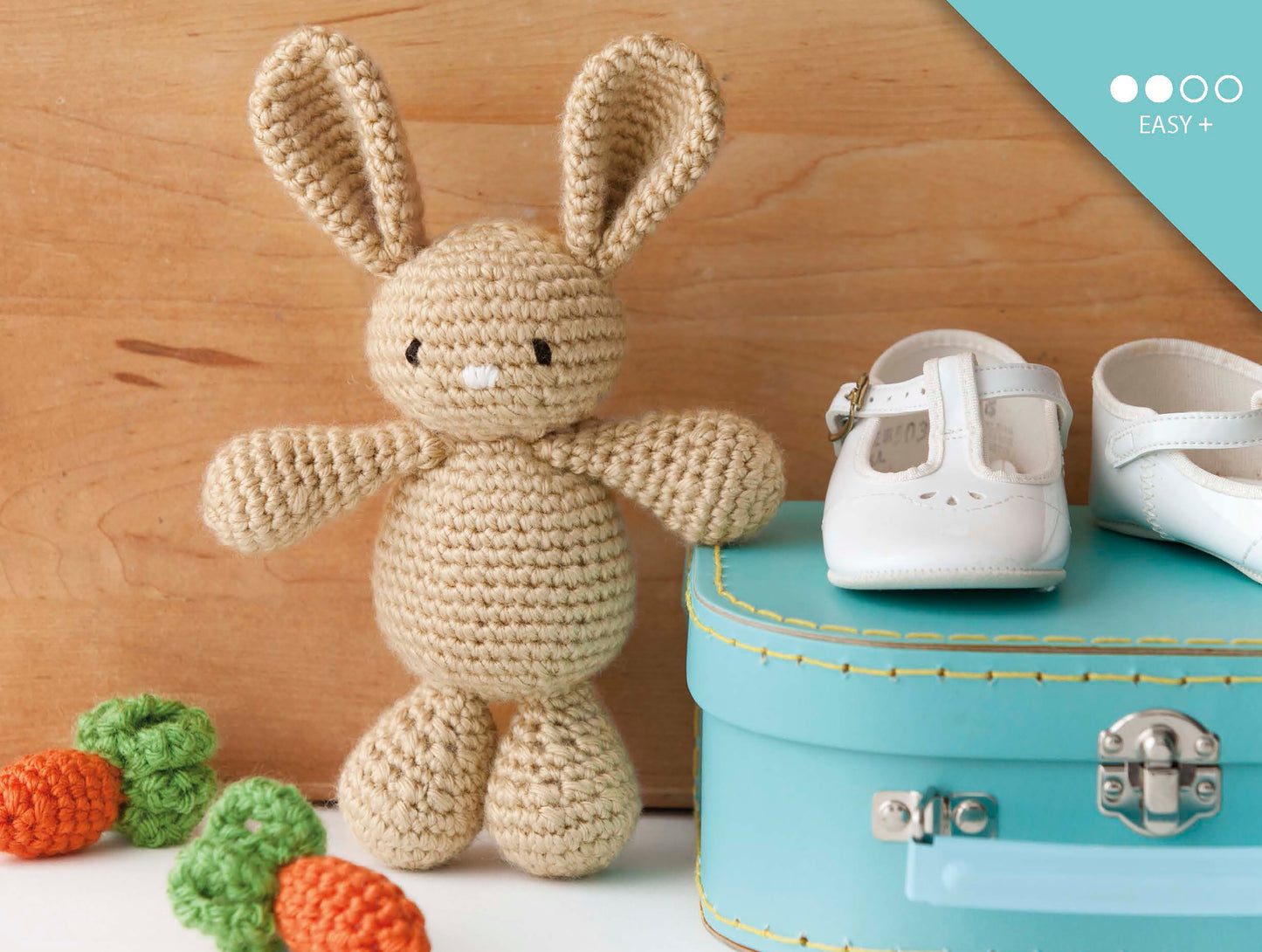 Leisure Arts' Baby's Buddy Amigurumi, a crocheted beige bunny made from beginner patterns, sits next to a turquoise suitcase topped with white baby shoes; crocheted carrots and peas accent the wooden surface.