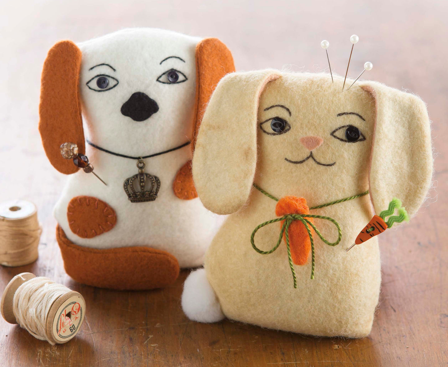 Two handmade felt dog plushies from the “My Pet Crafts” kit by Leisure Arts sit side by side, featuring embroidered faces and cute accessories. One holds a carrot, and both have spools of thread nearby on a wooden surface—a delightful showcase of animal sewing patterns, perfect for pet lover gifts.