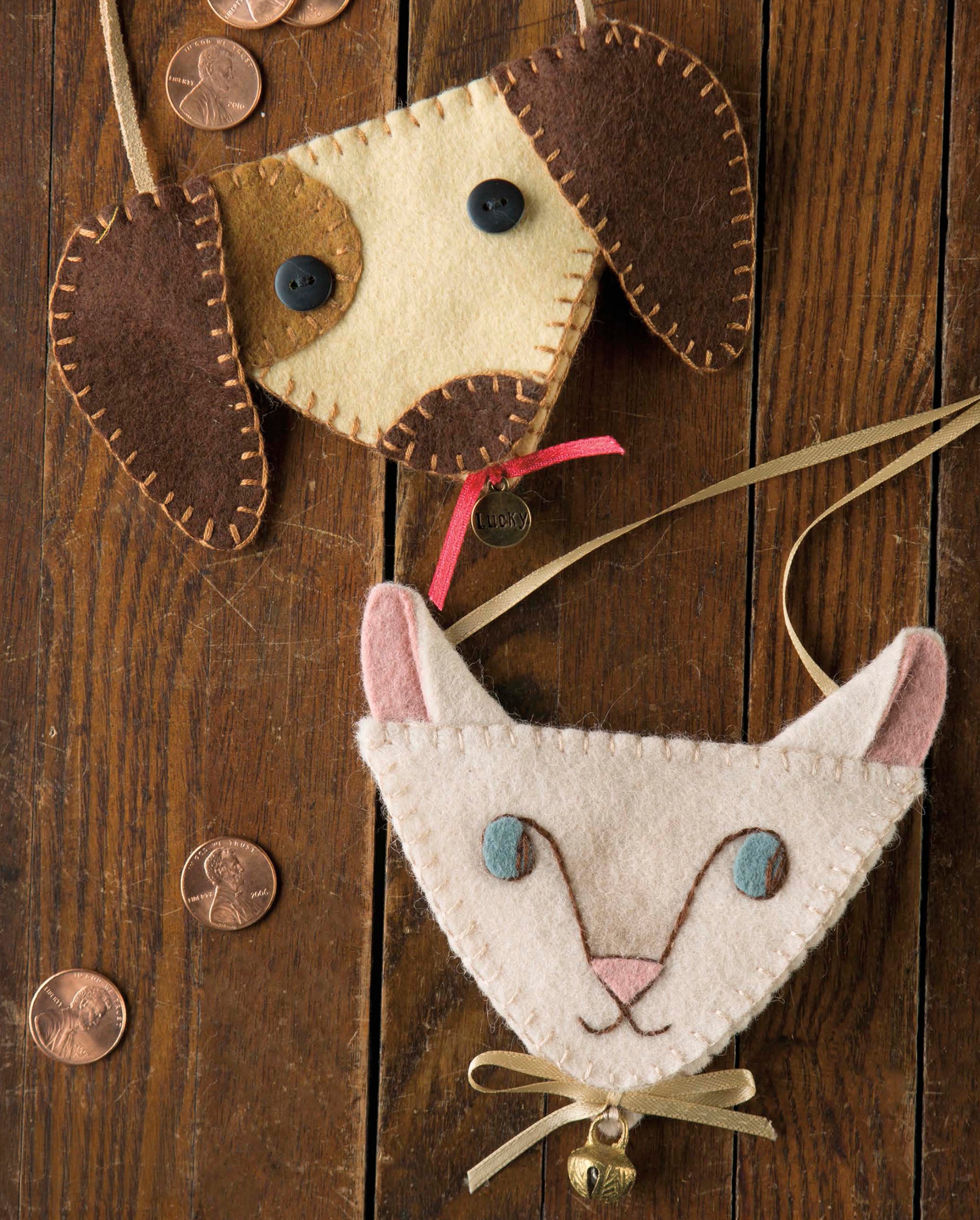 Two handmade felt pouches from the Leisure Arts My Pet Crafts collection, shaped like a dog and a cat, rest on a wooden surface scattered with pennies. The dog features button eyes, while the cat sports a stitched face and a bell hanging from its ribbon—delightful handcrafted pieces that make perfect gifts for pet lovers.