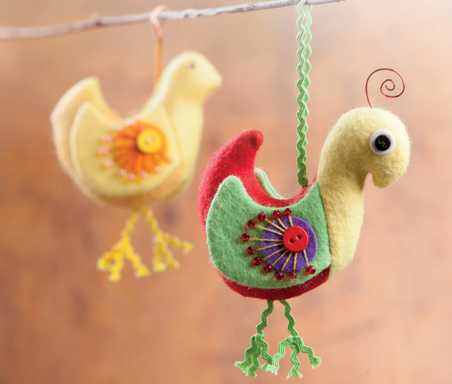 Two vibrant felt bird ornaments from Leisure Arts’ My Pet Crafts collection hang gracefully from a branch, adorned with charming button and bead accents. With one bird in sharp focus and the other gently blurred in the background, these delightful handmade creations make wonderful gifts for pet lovers or whimsical touches inspired by animal sewing patterns.