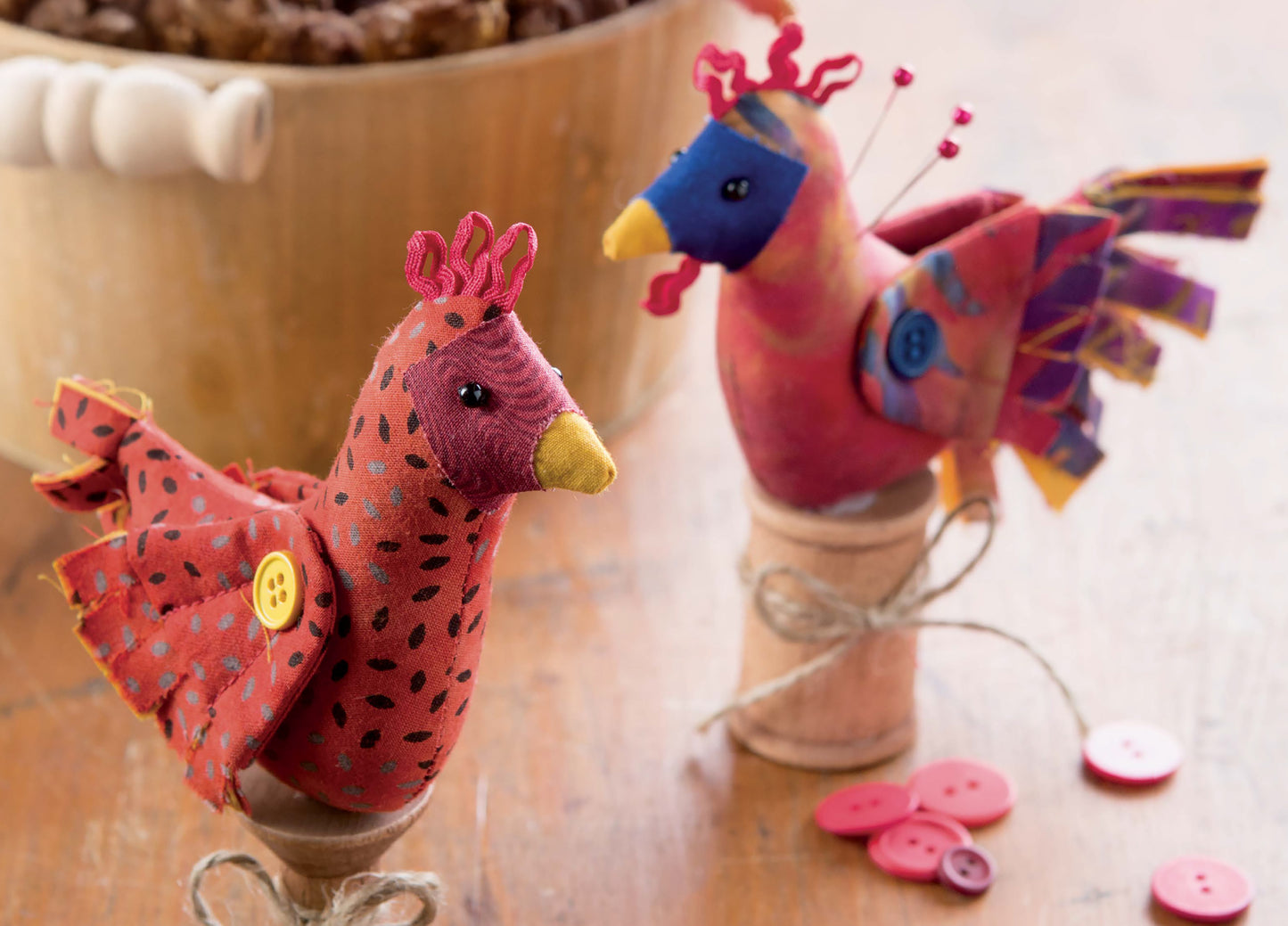 Two vibrant, handmade fabric birds from My Pet Crafts by Leisure Arts, featuring button wings and twine-wrapped wooden spool bases, are displayed on a wooden surface. Ideal for enthusiasts of handmade pet crafts, pink buttons are scattered nearby and a wooden bowl sits in the background.