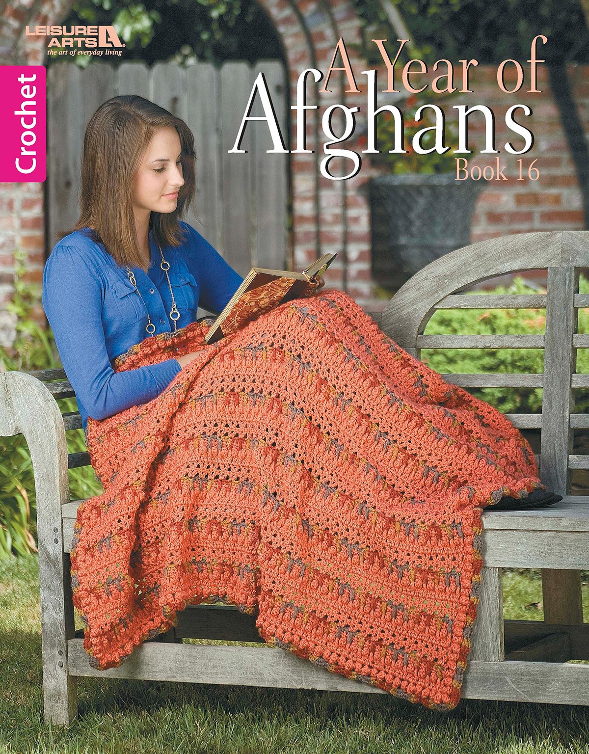 A woman crochets an orange afghan with medium weight yarn outdoors on a wooden bench. The digital download, "A Year of Afghans, Book 16" by Leisure Arts, is pictured beside her in a garden surrounded by lush greenery and a fence.