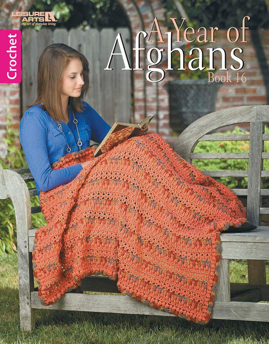 A woman crochets an orange afghan with medium weight yarn outdoors on a wooden bench. The digital download, "A Year of Afghans, Book 16" by Leisure Arts, is pictured beside her in a garden surrounded by lush greenery and a fence.