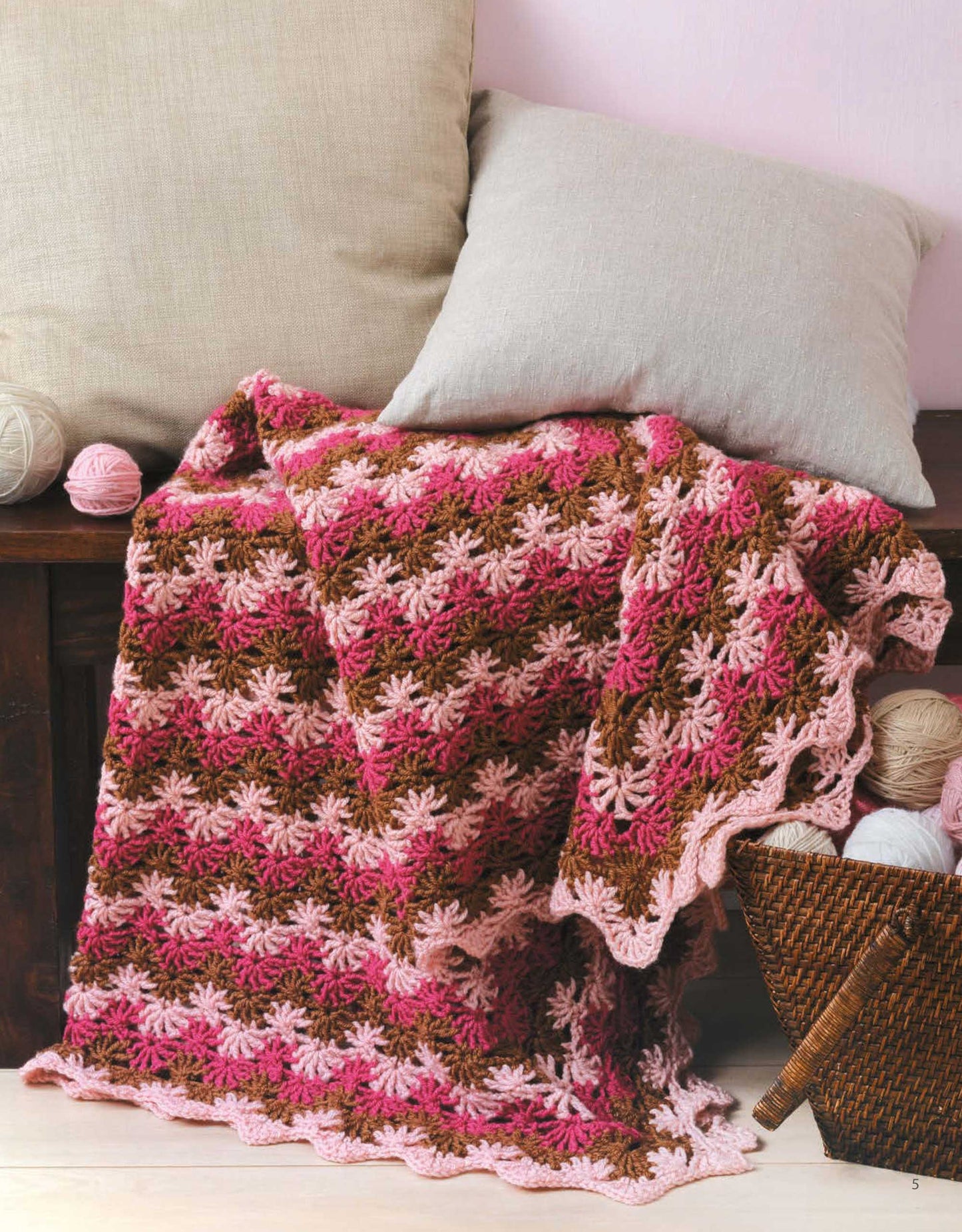 The "A Year of Afghans, Book 16 Digital Download" by Leisure Arts features crocheted blankets in pink, brown, and cream using medium weight yarn, shown on a dark wooden bench beside a wicker basket with colorful yarn balls.