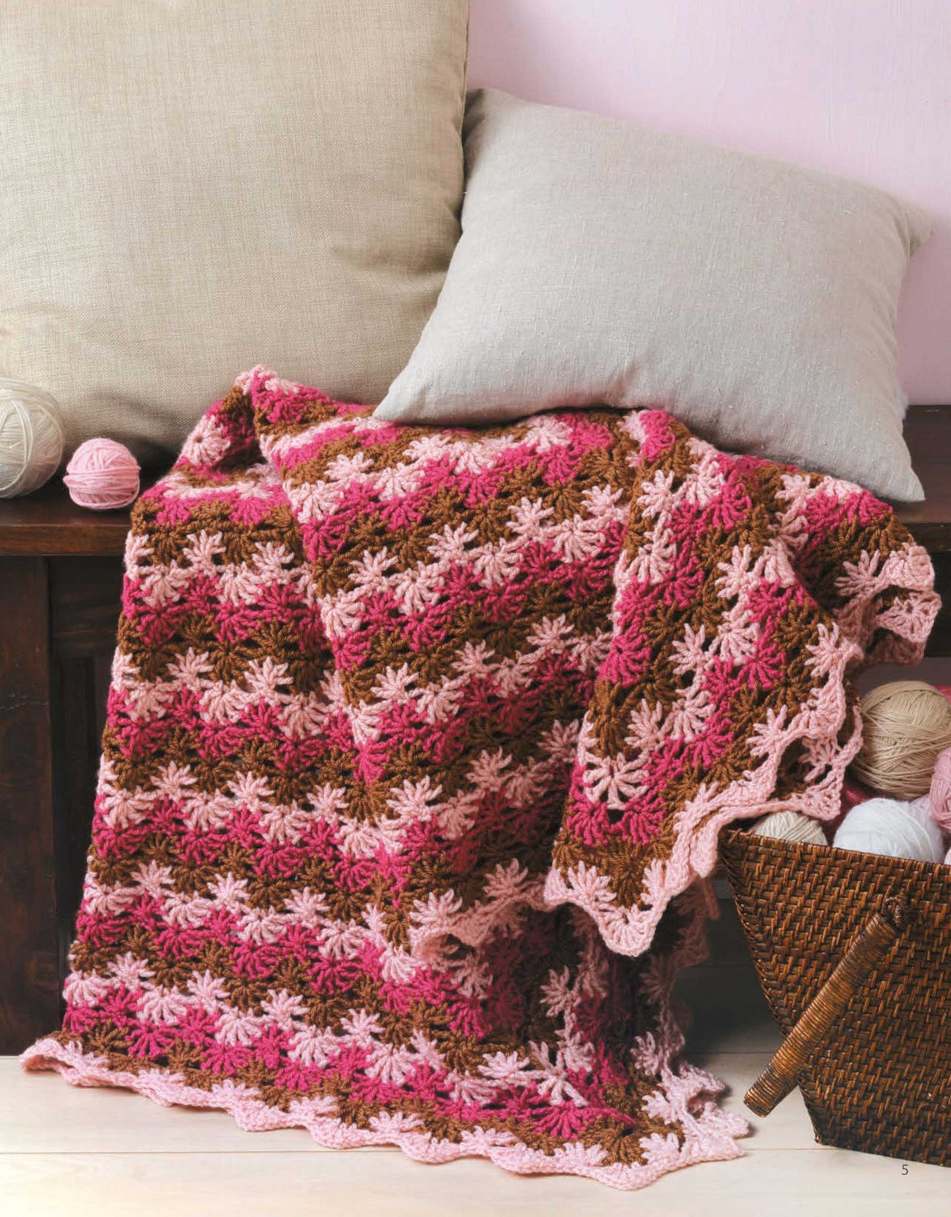 The "A Year of Afghans, Book 16 Digital Download" by Leisure Arts features crocheted blankets in pink, brown, and cream using medium weight yarn, shown on a dark wooden bench beside a wicker basket with colorful yarn balls.