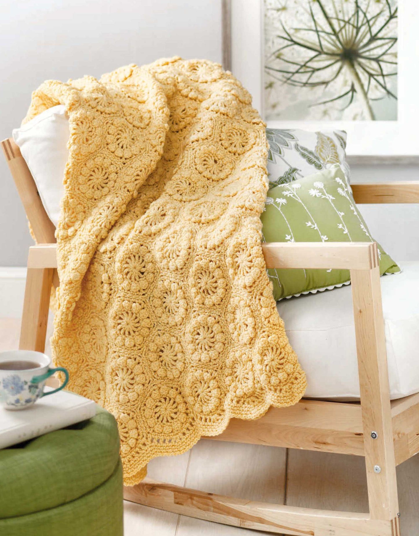 A cozy nook features a crocheted blanket from A Year of Afghans, Book 16 by Leisure Arts draped on a wooden chair with green and white cushions. Nearby, a cup of tea and botanical art complete the inviting scene.