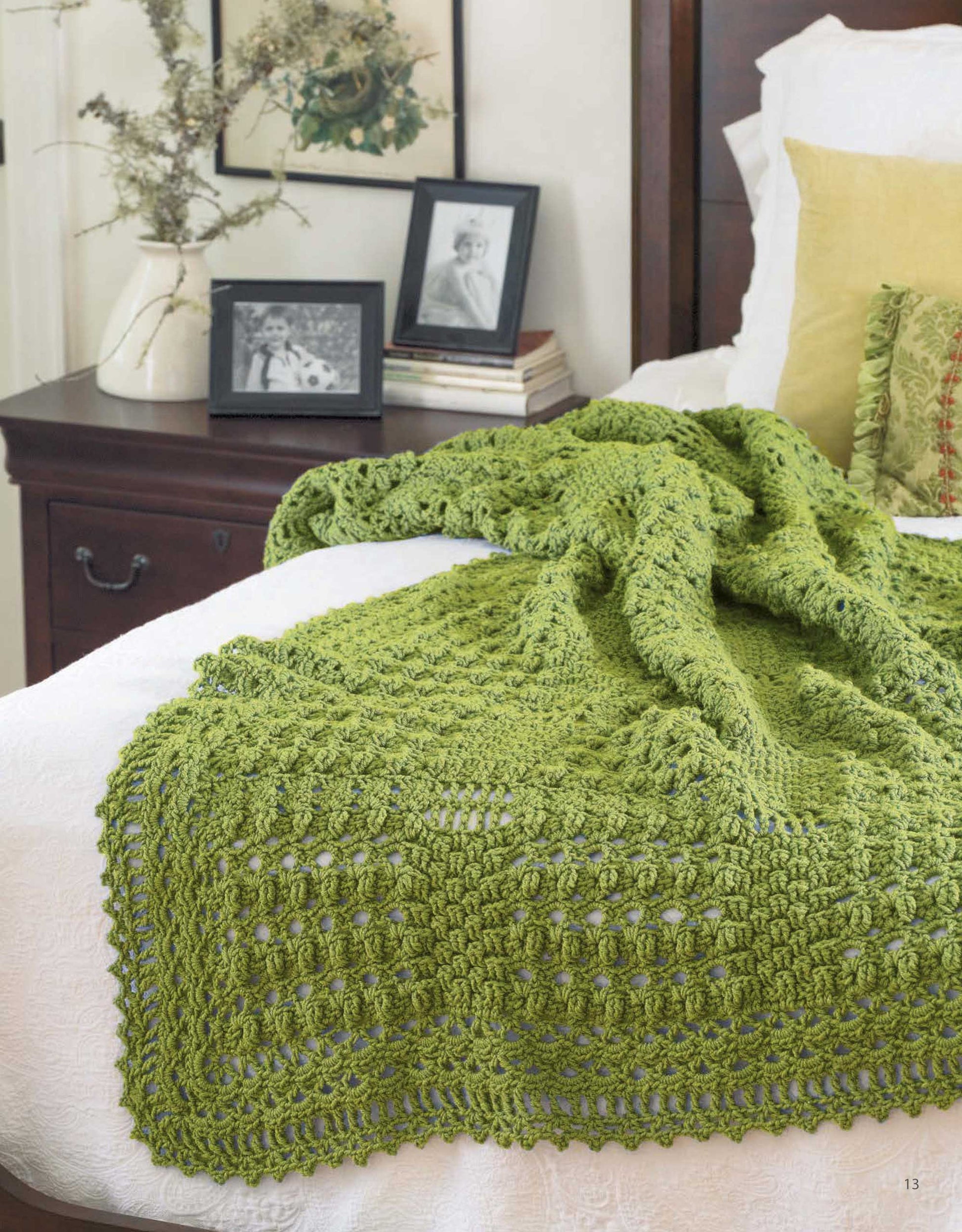 The Leisure Arts "A Year of Afghans, Book 16" digital download features a green crocheted blanket made with medium weight yarn, shown on a white bedspread beside framed photos, dried flowers, and a green pillow.