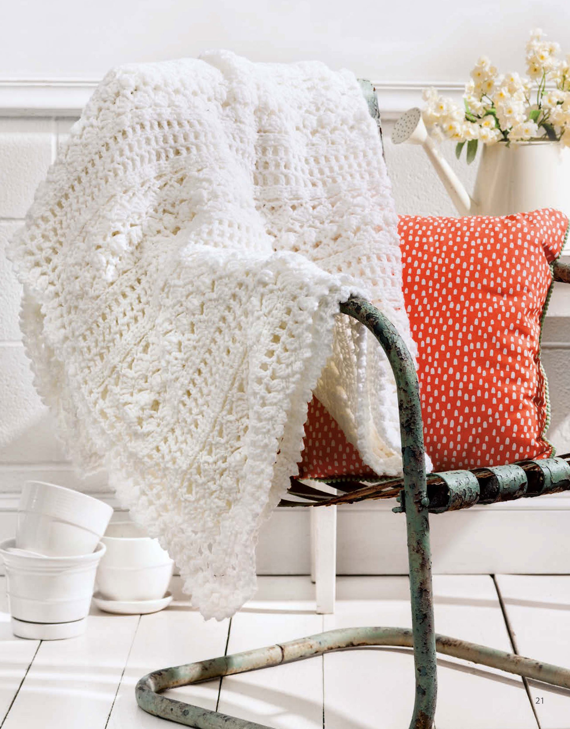 The Leisure Arts "A Year of Afghans, Book 16" digital download features a white crocheted blanket made with medium weight yarn, draped over a vintage metal chair beside a red patterned cushion and bright decor.