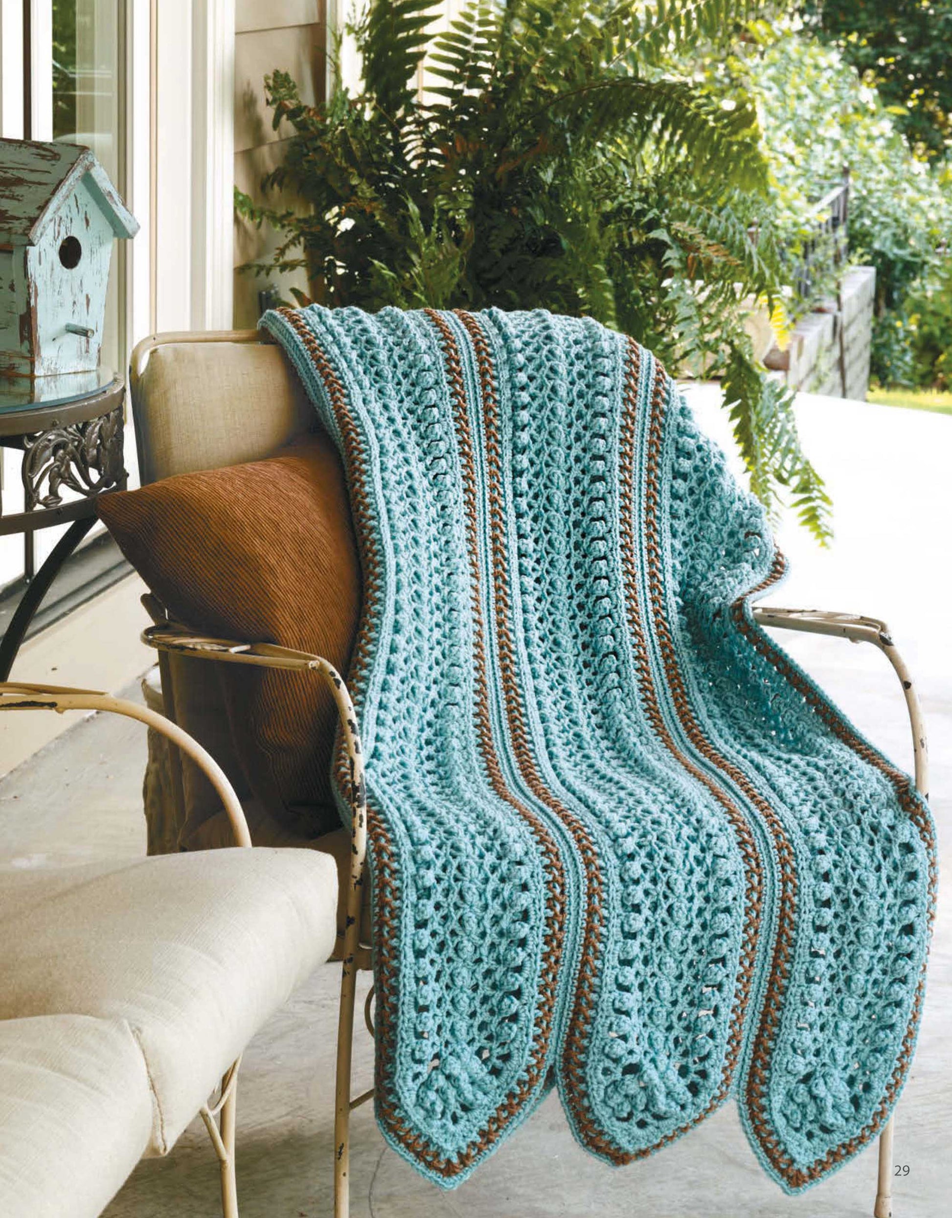 A striped blue and brown afghan from "A Year of Afghans, Book 16" by Leisure Arts is draped over a cushioned metal porch chair with a brown pillow. Green plants and a birdhouse appear in the background.