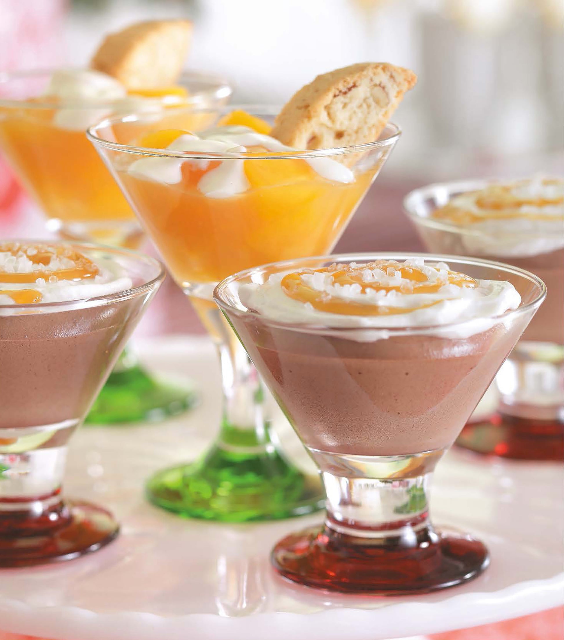 Open House Merriment by Leisure Arts features assorted dessert mousses elegantly served in martini glasses, topped with whipped cream, fruit, and biscotti cookies on a white plate—an ideal choice to add festive accents to any celebration.