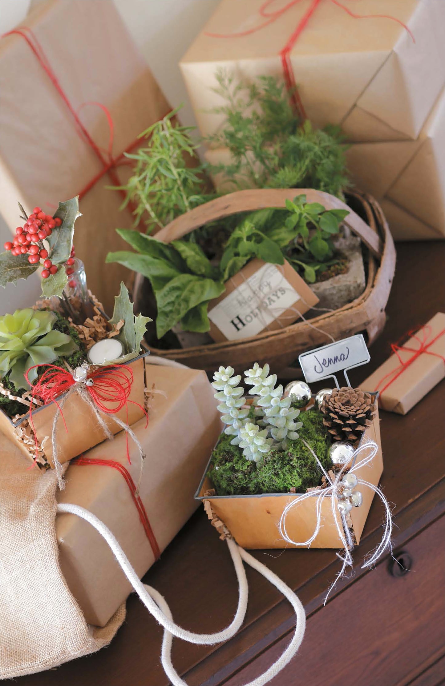 Open House Merriment by Leisure Arts displays gifts wrapped in brown paper and tied with red string, nestled beside potted plants, succulents, greenery, and festive accents on a wooden surface.