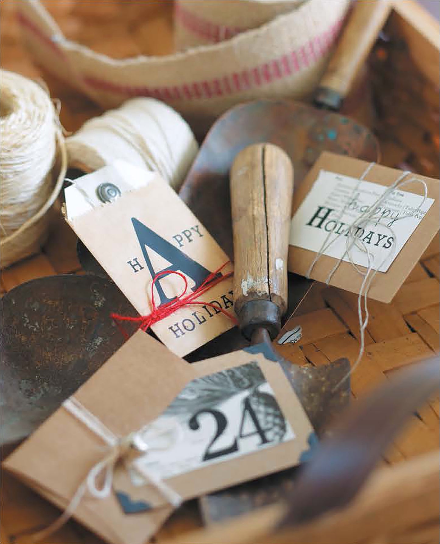 Open House Merriment gift tags by Leisure Arts, reading “Happy Holidays” and “24,” are nestled among old garden tools, twine, and ribbon in a woven basket, creating a rustic holiday arrangement with festive accents.
