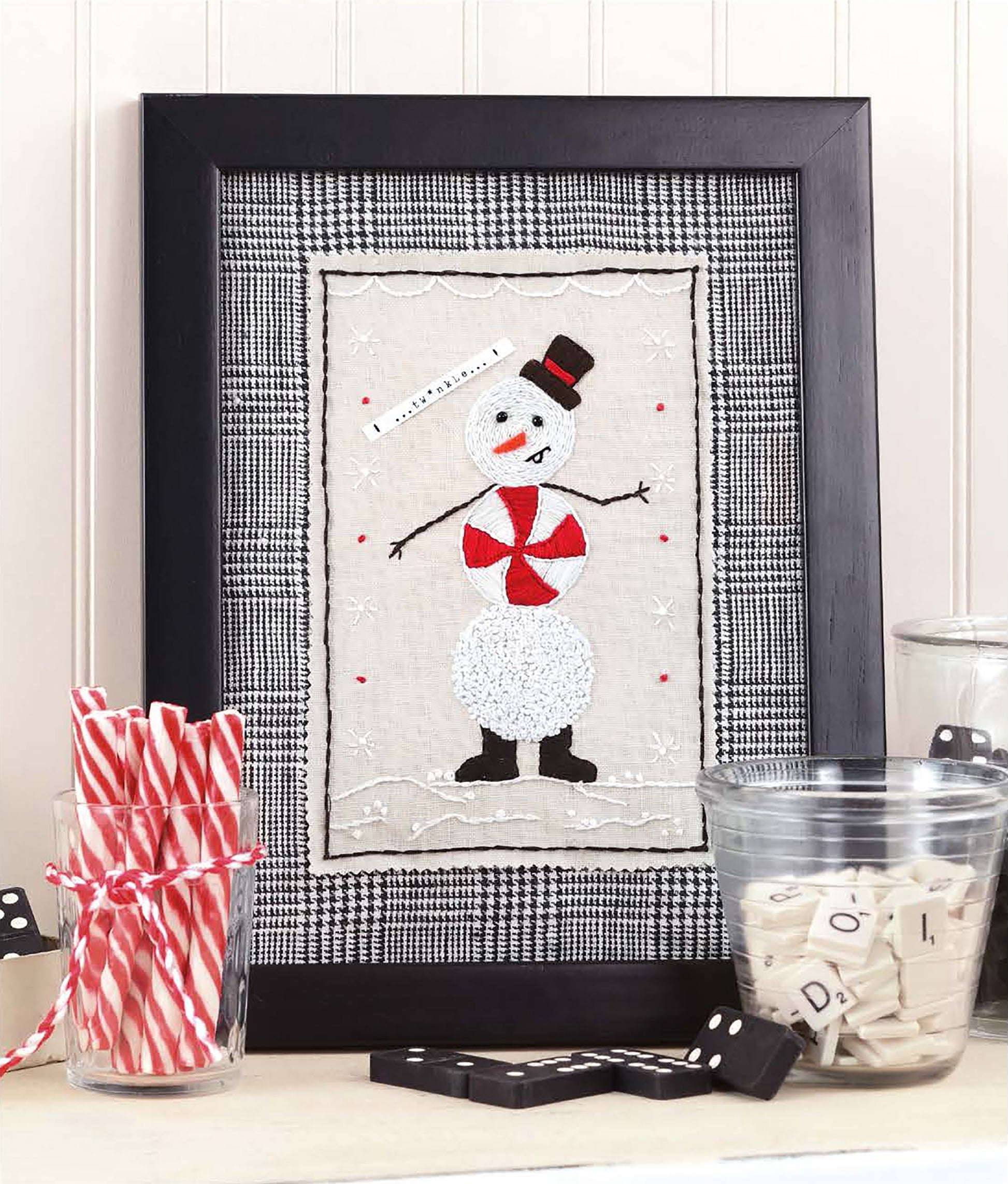 Open House Merriment by Leisure Arts features framed stitched snowman art accented with a red and white scarf, black hat, and boots. Surrounded by glasses filled with red-striped sticks and Scrabble tiles atop a checkered cloth, this piece is perfect for adding festive cheer to your holiday décor.
