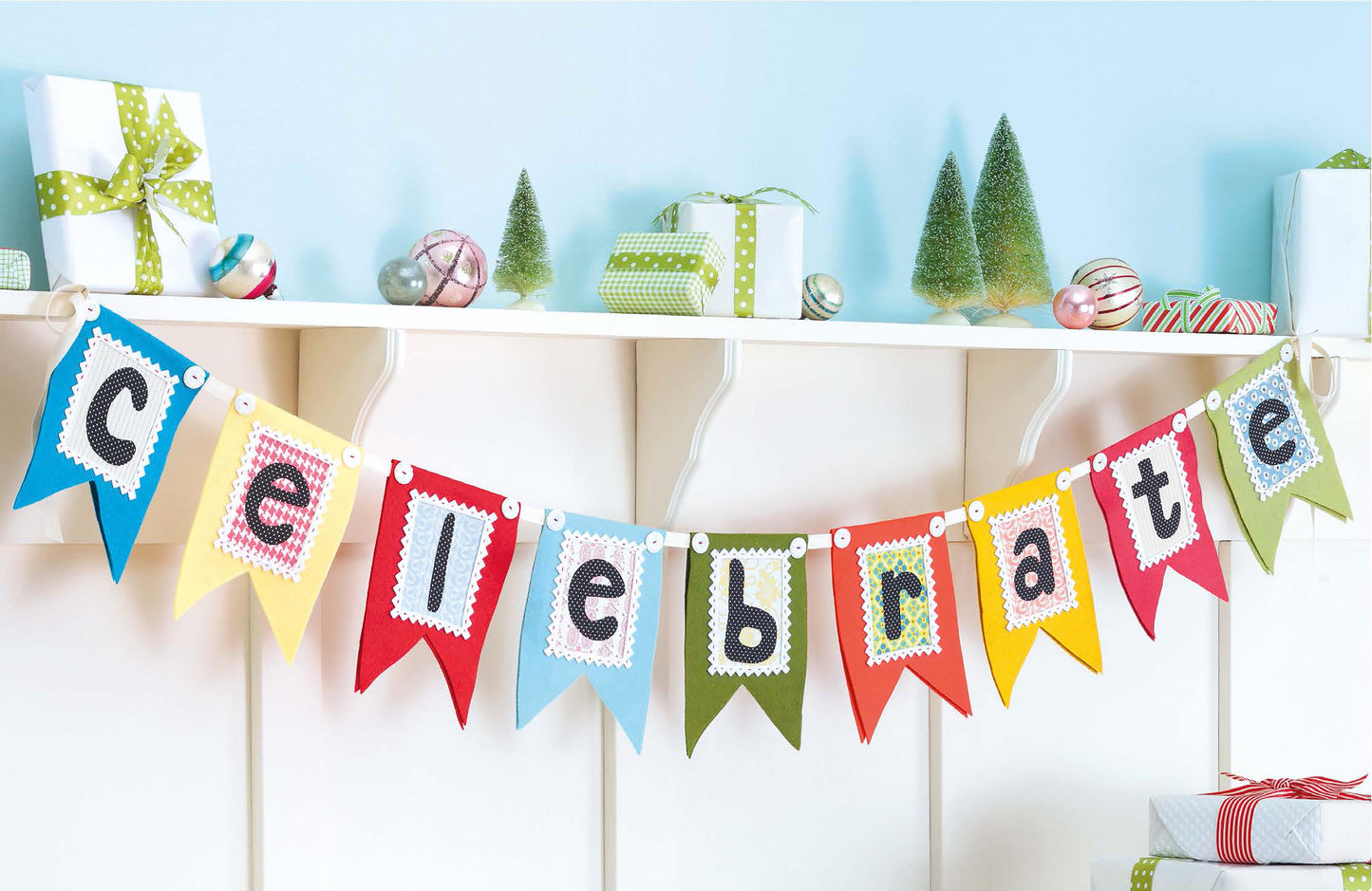 Add a festive touch to your space with Open House Merriment from Leisure Arts—a colorful pennant banner spelling “Celebrate” that hangs beneath a white shelf adorned with presents, ornaments, and small green Christmas trees against a light blue wall—perfect inspiration for quick and easy crafts this season.