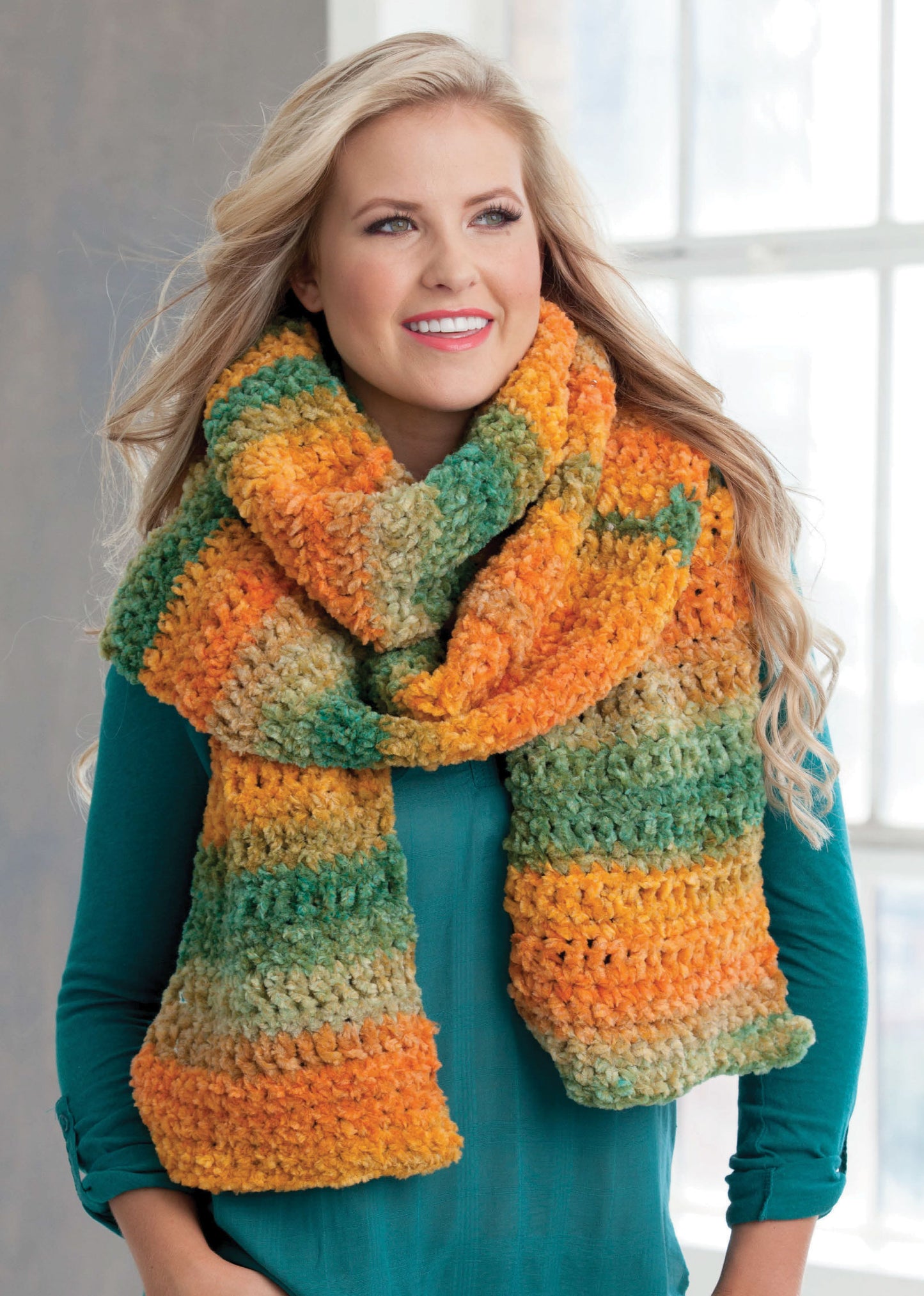 A woman with long blonde hair smiles indoors near a window, wearing a chunky, multicolored scarf made from beginner projects in orange, green, and yellow using the "Learn to Crochet the Easy Way" kit by Leisure Arts over a teal top.