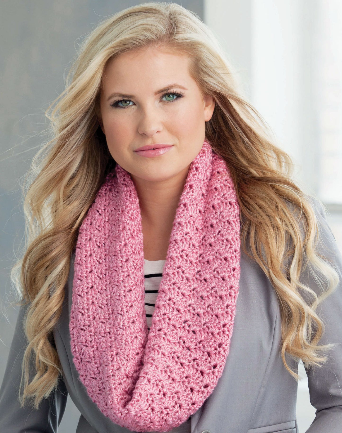 A woman with long blonde hair models a chunky pink infinity scarf from Leisure Arts' "Learn to Crochet the Easy Way," wearing it over a gray blazer and striped shirt, perfect for beginner crochet projects.