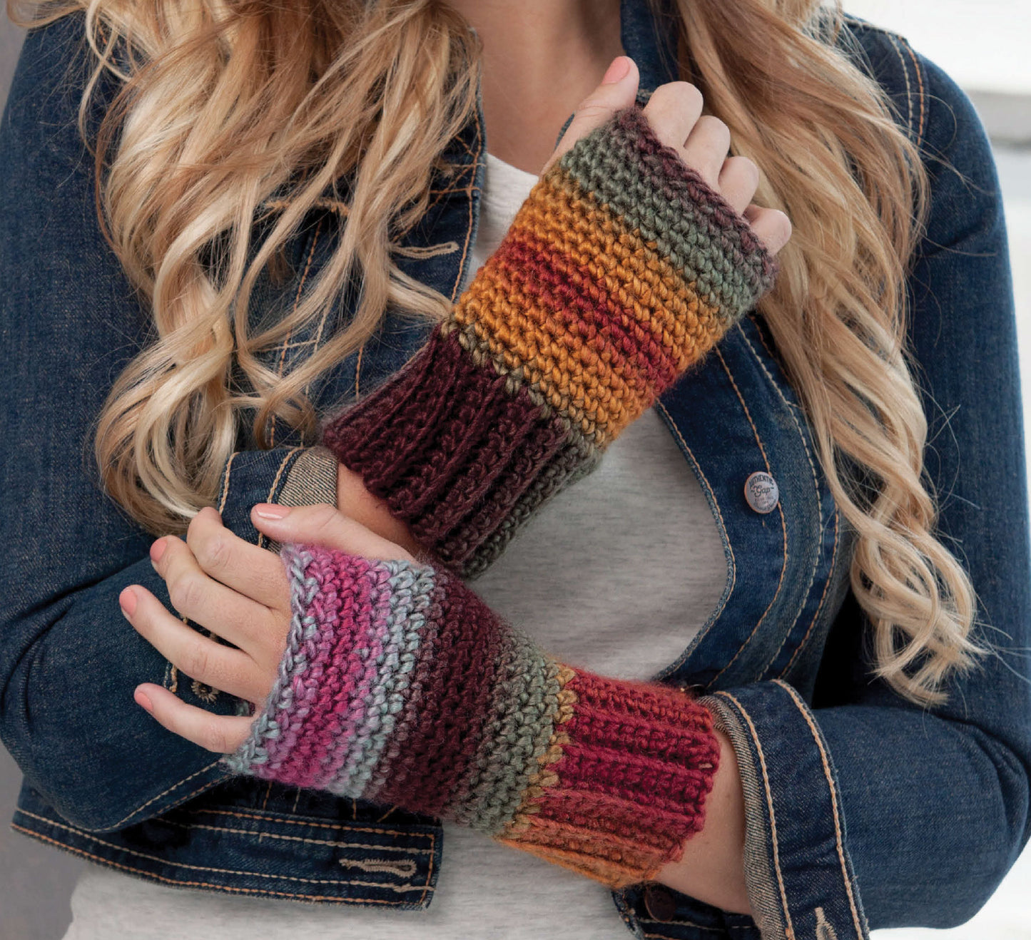 A person with long, wavy blonde hair wears a denim jacket and gray shirt, modeling colorful striped hand-knitted fingerless gloves—perfect inspiration from Leisure Arts' "Learn to Crochet the Easy Way" for beginner crochet projects.