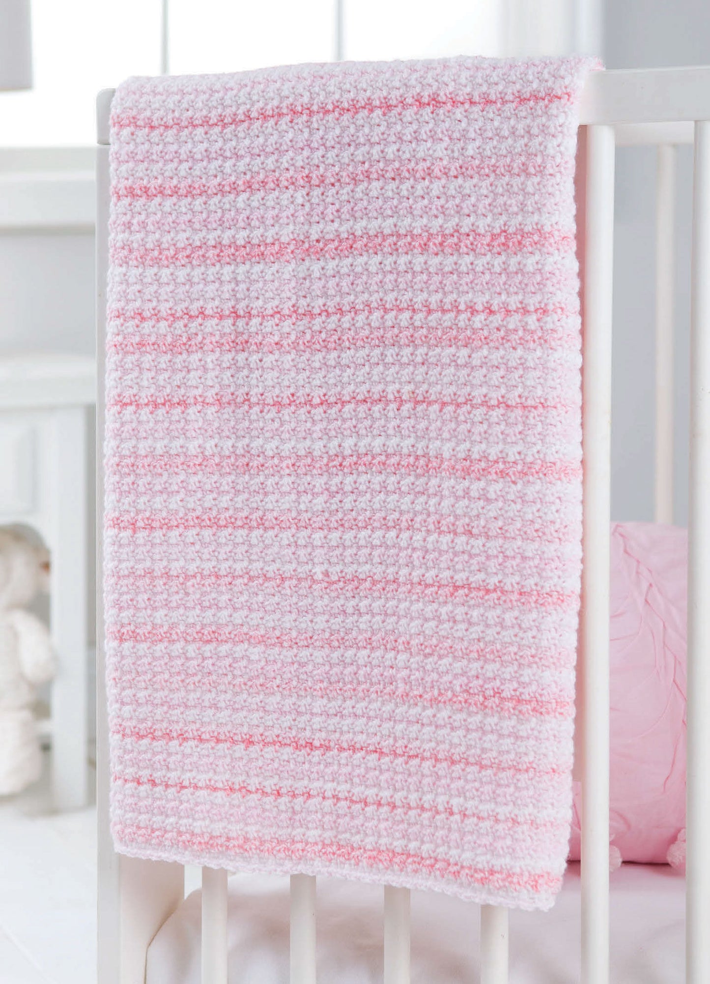 The Leisure Arts "Learn to Crochet the Easy Way" kit features a soft, textured pink and white striped baby blanket—ideal for beginners—shown draped over a white crib in a gently lit nursery.