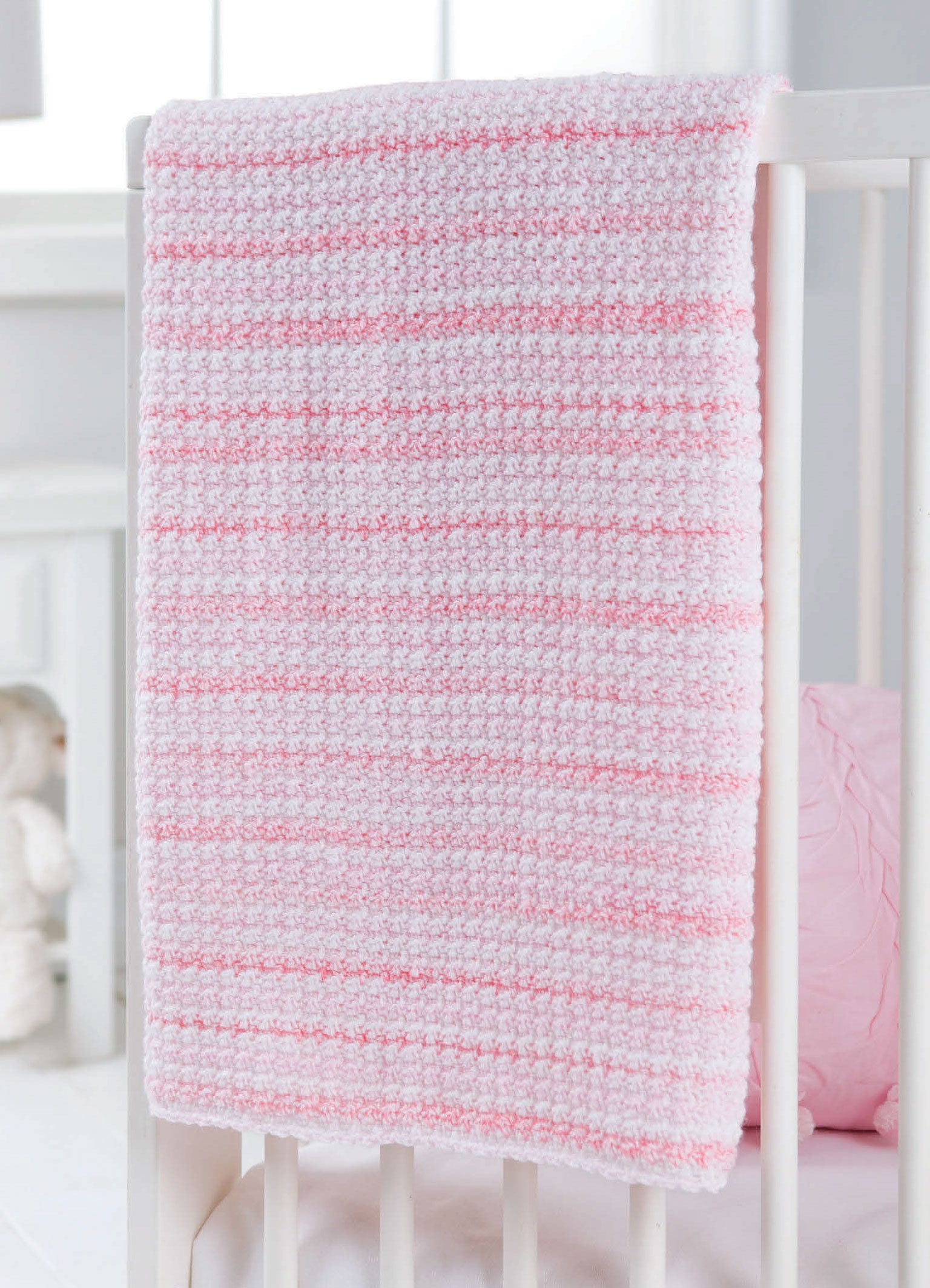 The Leisure Arts "Learn to Crochet the Easy Way" kit features a soft, textured pink and white striped baby blanket—ideal for beginners—shown draped over a white crib in a gently lit nursery.