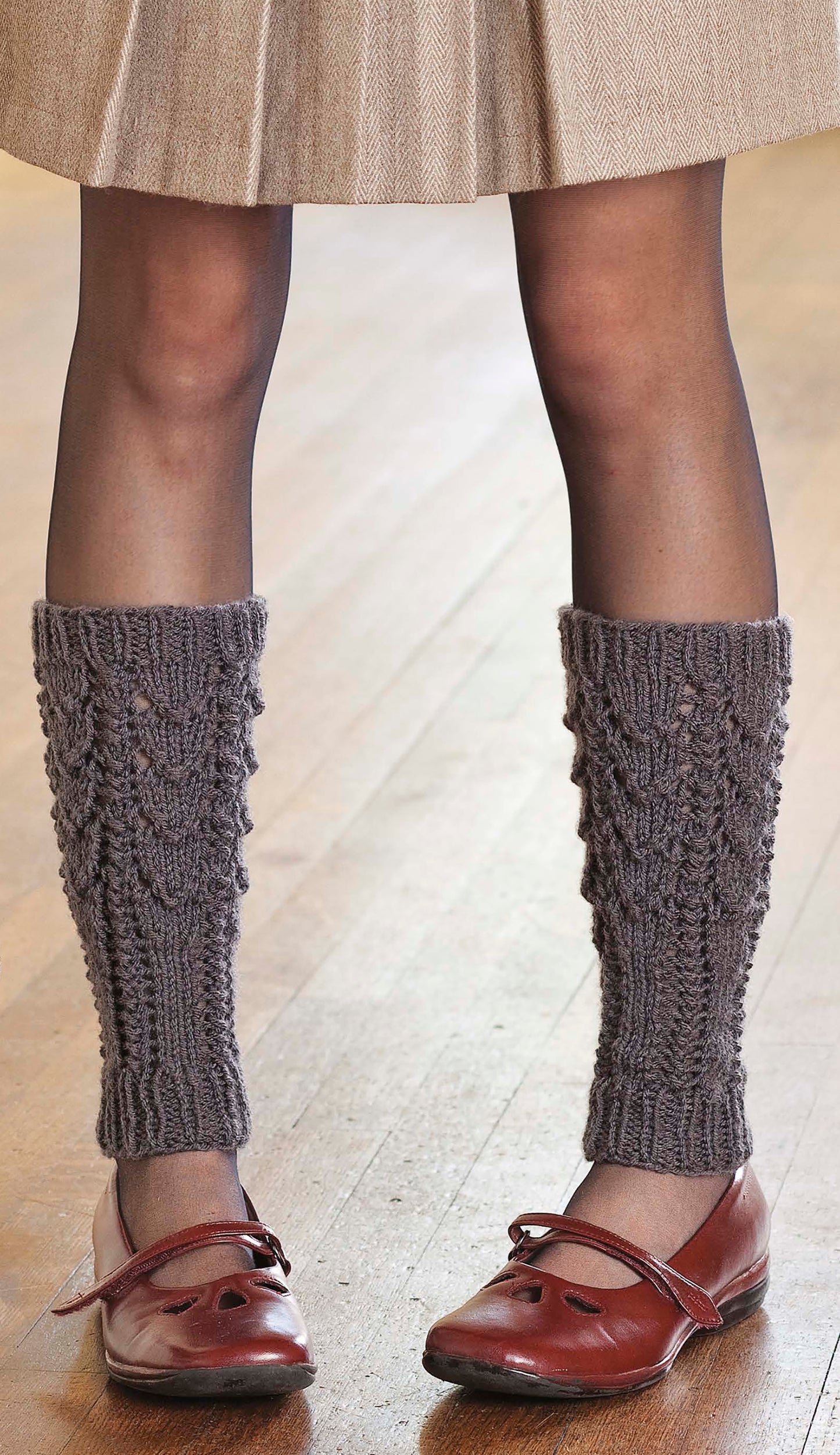 A person wears a beige skirt, dark sheer tights, red Mary Jane shoes, and gray knitted leg warmers made with Cozy Knits Made with the Knook by Leisure Arts while standing on a wooden floor.