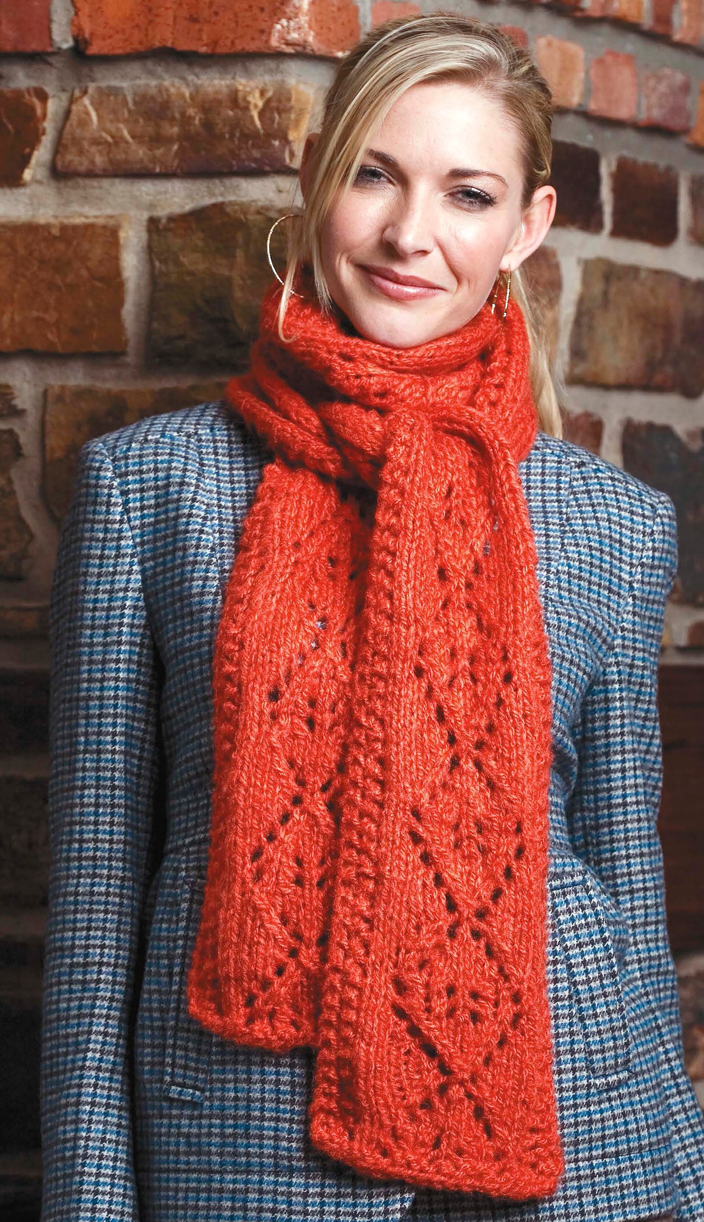 A woman wearing a gray plaid coat and a textured, bright red scarf made with Cozy Knits Made with the Knook by Leisure Arts stands in front of a brick wall, smiling at the camera.
