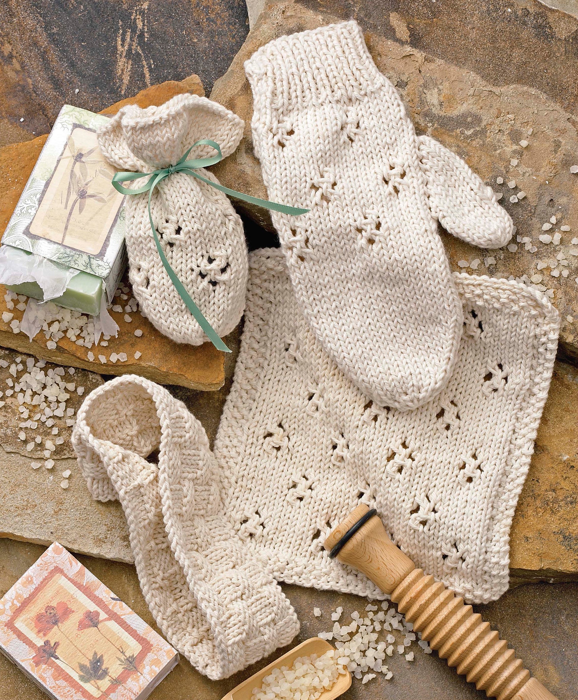 The Leisure Arts Cozy Knits Made with the Knook set features cream-colored spa accessories—a mitten, washcloth, headband, and drawstring pouch—beautifully displayed with soap bars, bath salts, and a wooden brush.