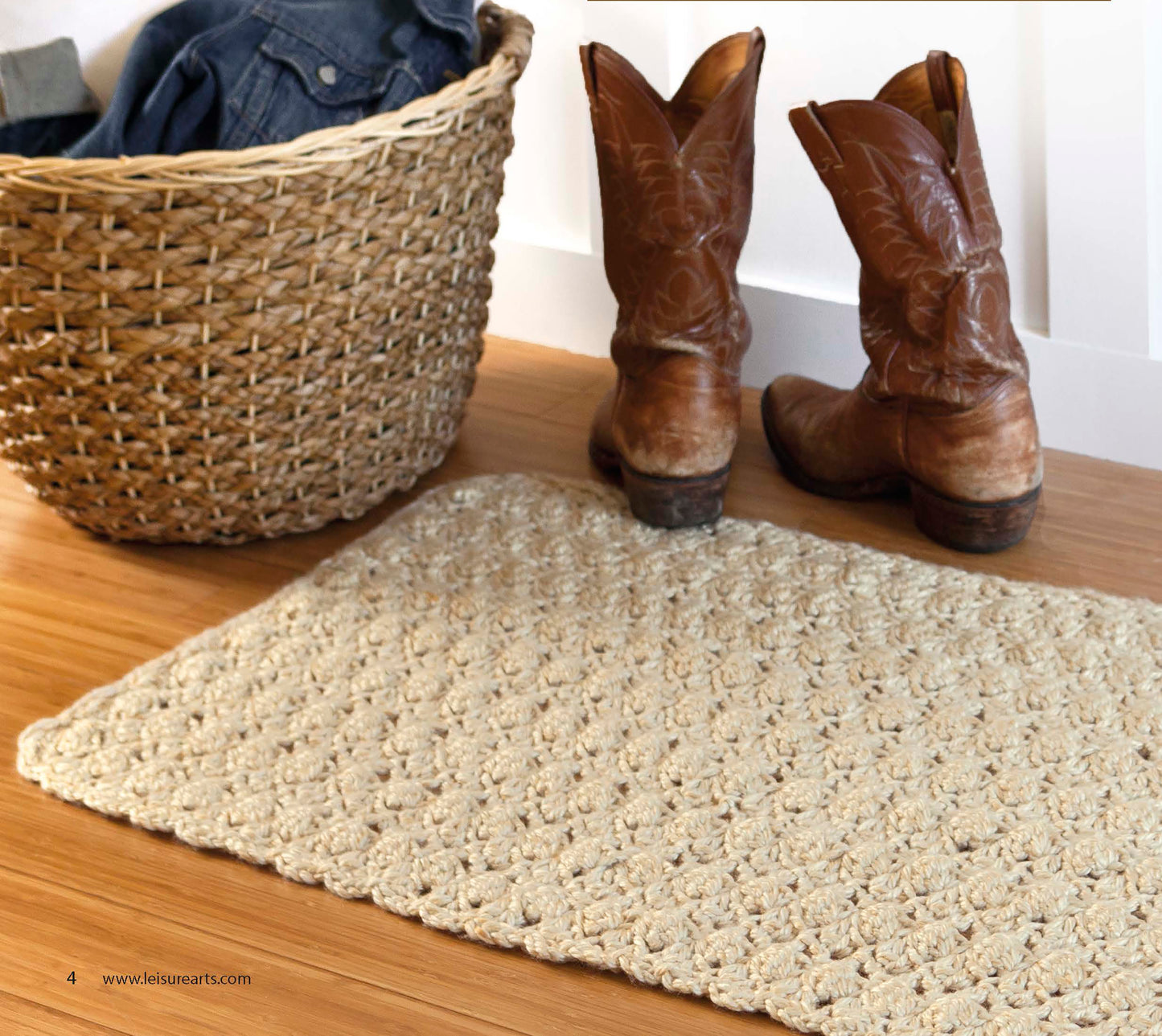 A pair of brown cowboy boots sits on a wooden floor beside a woven basket with blue clothing and a textured beige rug—perfect inspiration for making cozy crochet rugs from "Retro Rugs—Crochet 7 Quick Rugs" by Leisure Arts.