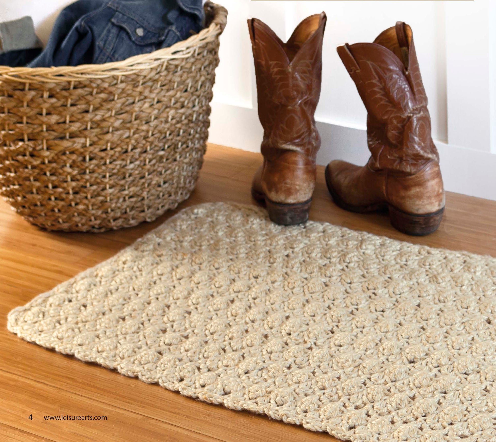 A pair of brown cowboy boots sits on a wooden floor beside a woven basket with blue clothing and a textured beige rug—perfect inspiration for making cozy crochet rugs from "Retro Rugs—Crochet 7 Quick Rugs" by Leisure Arts.