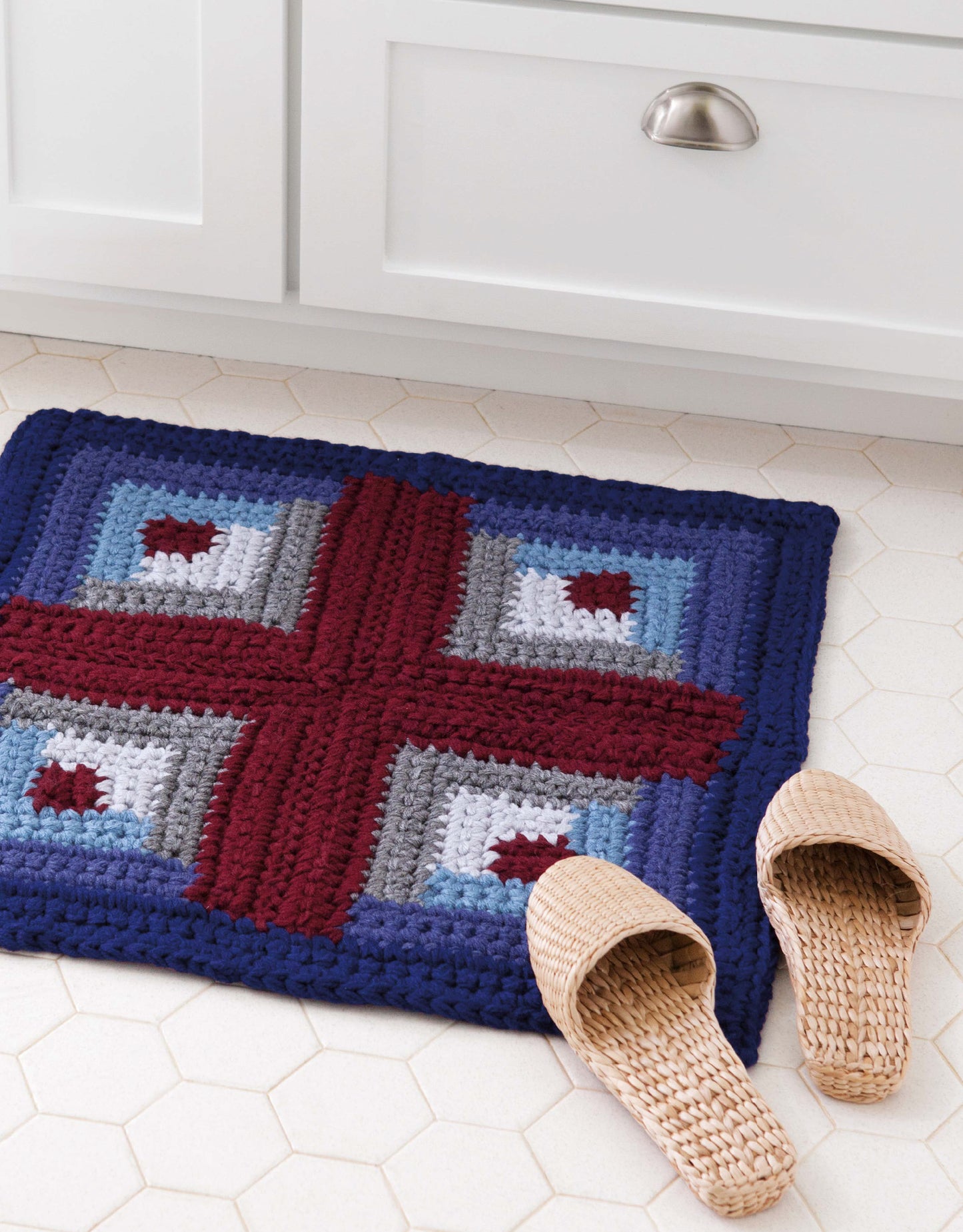 Get inspired by Retro Rugs—Crochet 7 Quick Rugs from Leisure Arts: a blue, gray, and burgundy geometric woven mat on white hexagonal tile floors beside slippers and a cabinet—perfect for beginner crochet or cozy rugs with super bulky yarn.