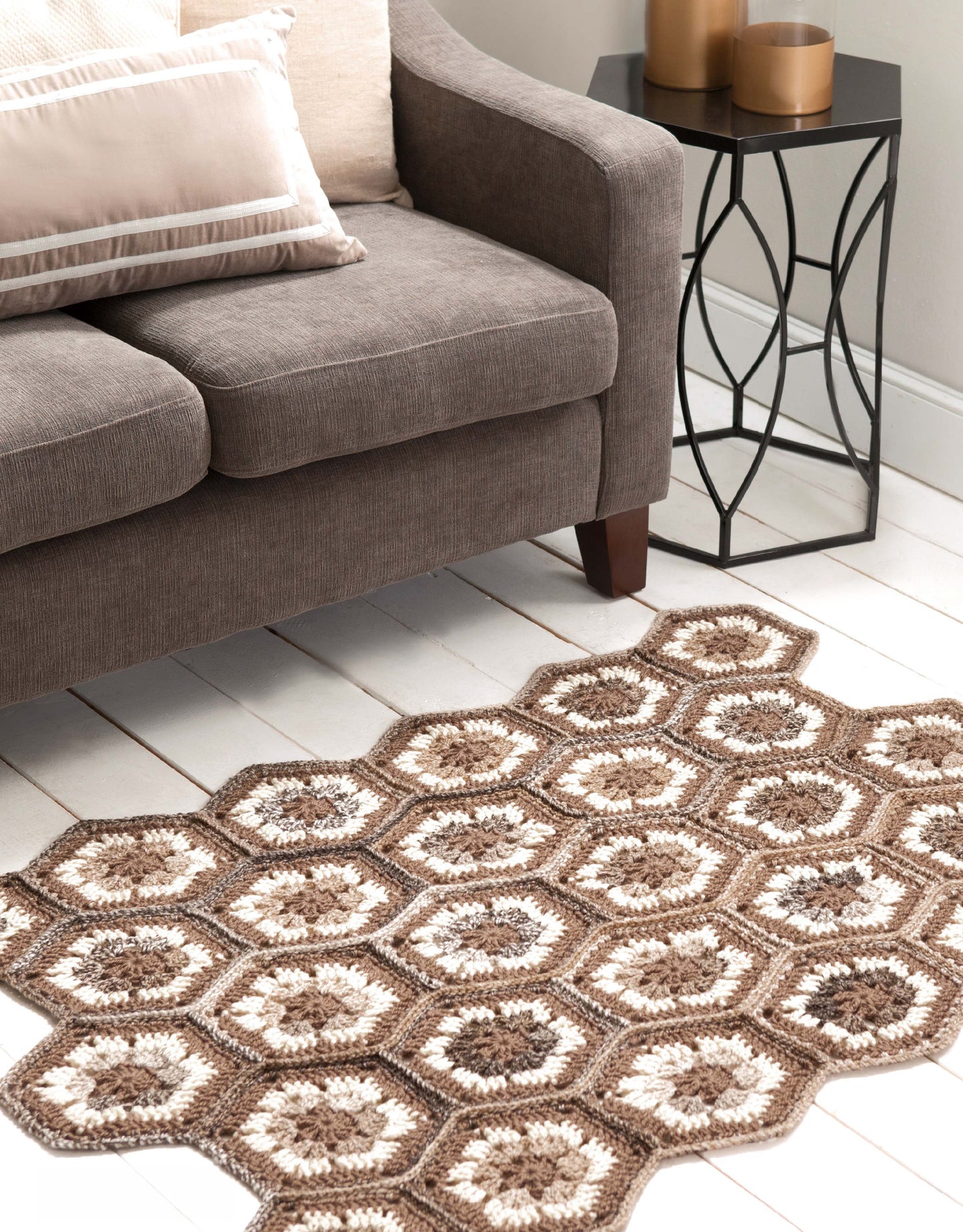 A cozy living room with a brown sofa, cream and beige pillows, a black side table topped with gold candles, and a hexagon-patterned Retro Rugs—Crochet 7 Quick Rugs by Leisure Arts in brown and cream on a white wooden floor.