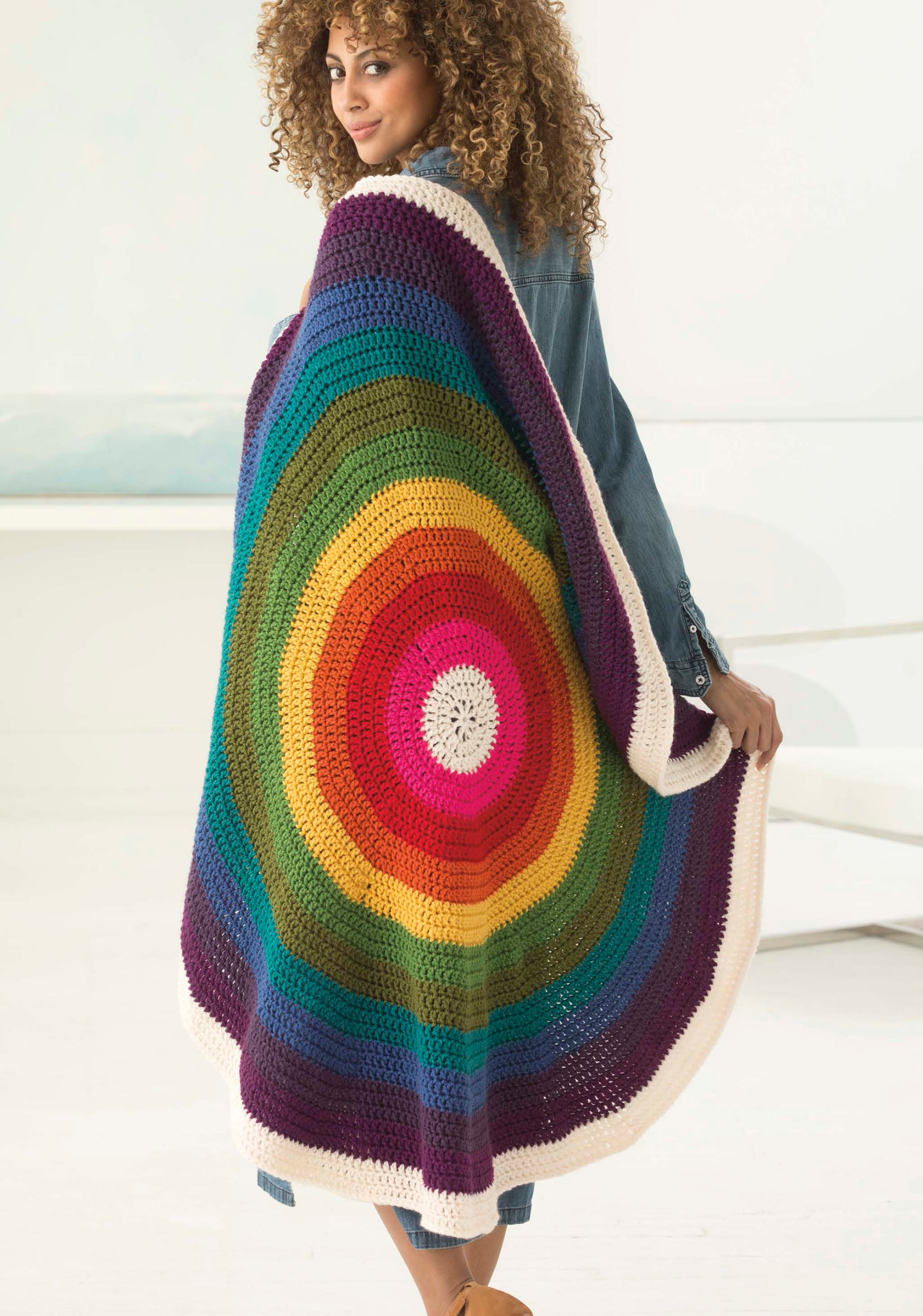 A woman with curly hair drapes the Leisure Arts Mindful Mandala Afghan, featuring vibrant concentric rainbow circles and a white border, over her shoulders as she stands in a bright, modern room.