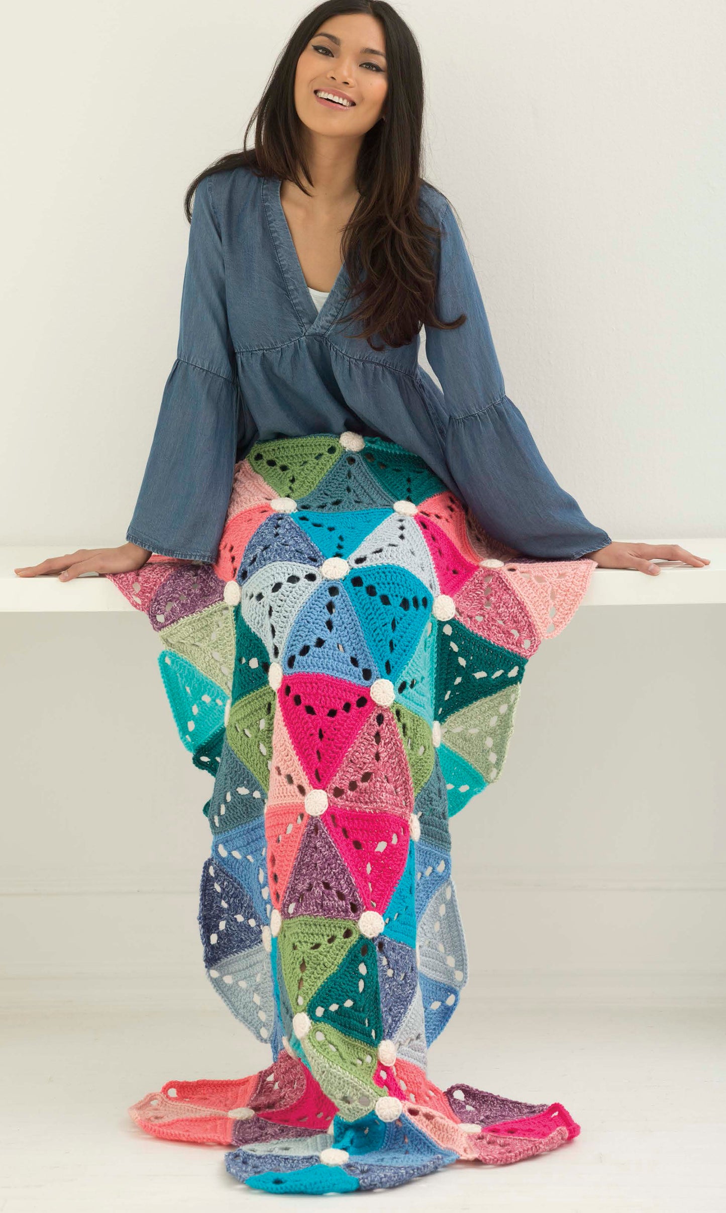 A smiling woman sits on a white bench, wearing a blue blouse and wrapped in the Mindful Mandala Afghans by Leisure Arts, featuring colorful, intricate crochet patterns draped over her lap and flowing gracefully to the floor.