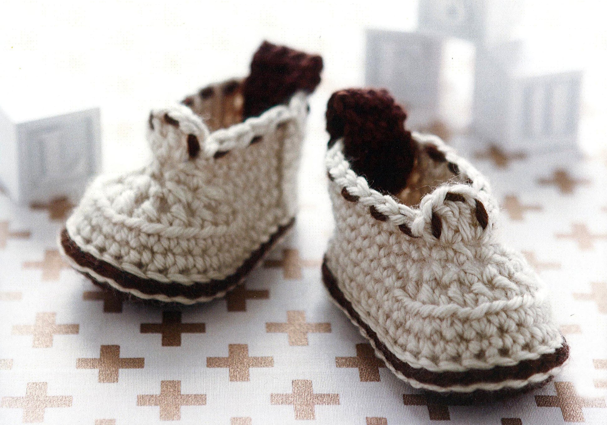 Two cream baby booties with brown accents from Leisure Arts’ Modern Baby Booties Digital Download rest on a patterned surface. Great for those seeking beginner crochet patterns for baby shoes. Blurred white toy blocks are in the background.
