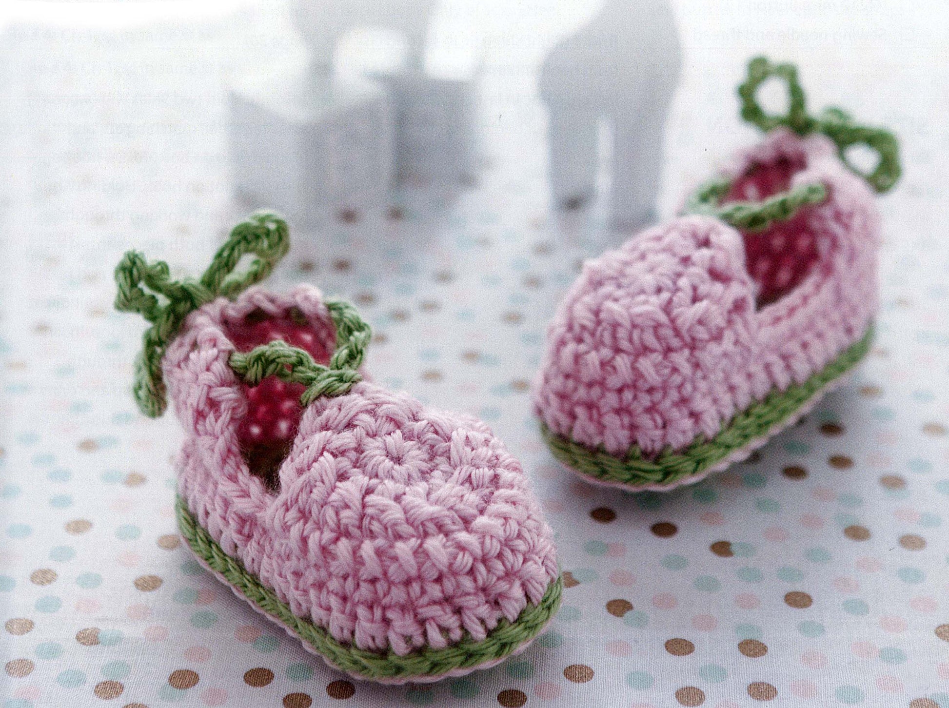 Two small pink and green crochet baby booties, made with beginner patterns from the Modern Baby Booties Digital Download by Leisure Arts, rest on a white surface with pastel polka dots. Blurry animal-shaped toys appear in the background.