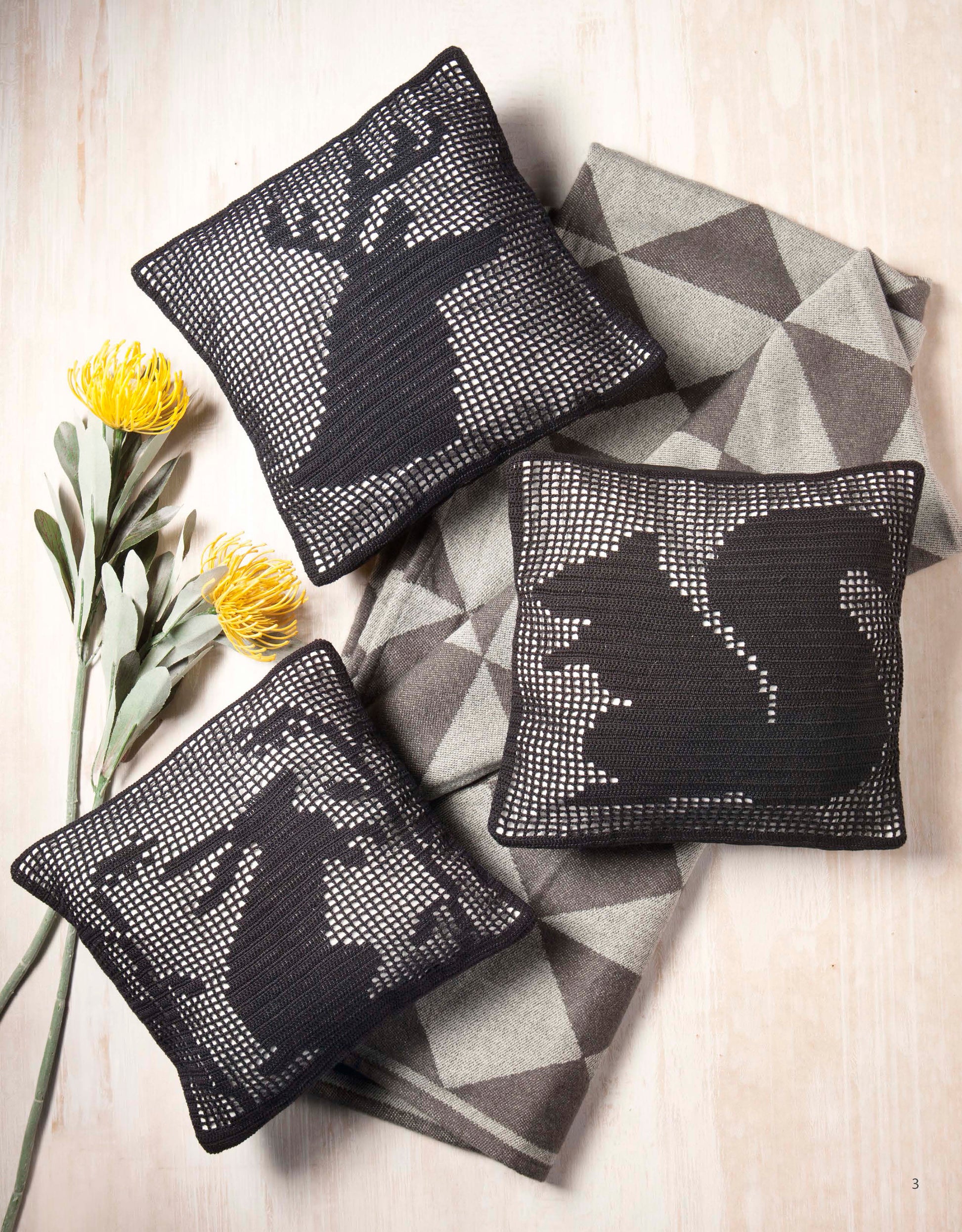 Three black and white "Woodland Wildlife in Filet Crochet" cushions by Leisure Arts, featuring deer, hare, and squirrel silhouettes, rest on a geometric blanket beside two yellow flowers on light wood.