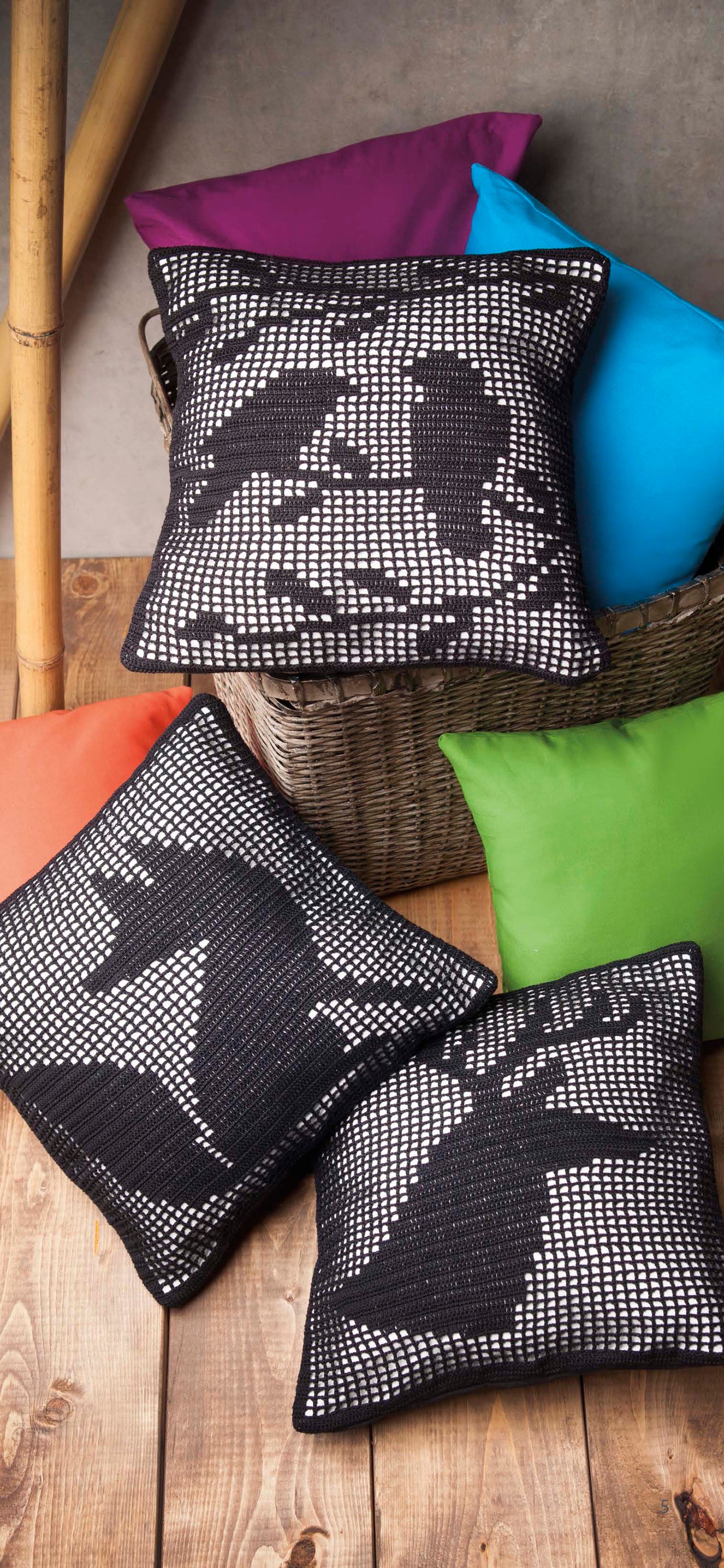 Three "Woodland Wildlife in Filet Crochet" cushions by Leisure Arts, featuring black and white animal crochet designs, are displayed on a wooden floor and in a wicker basket among plain purple, blue, and green cushions with bamboo stalks behind.