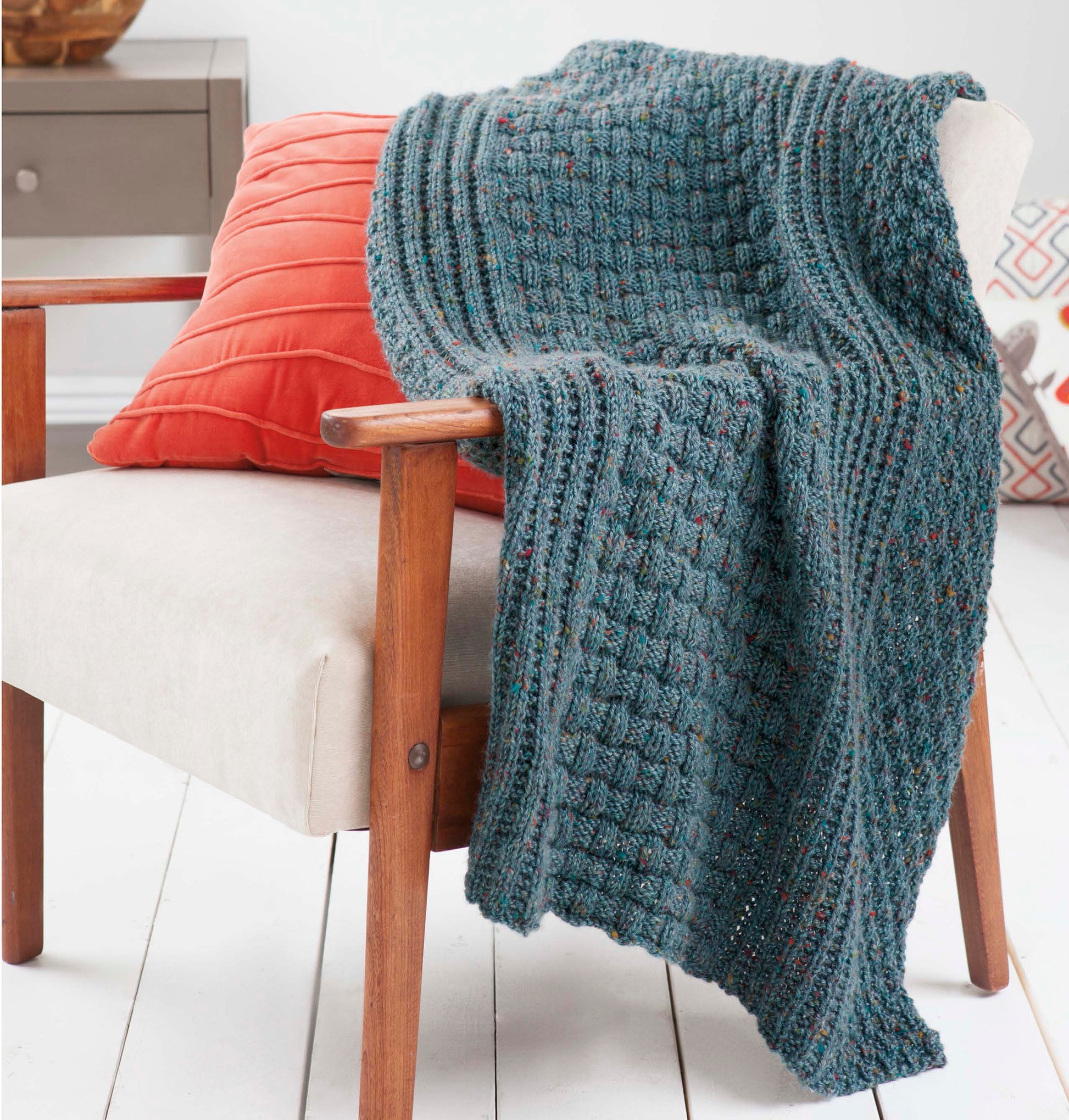 The Loom Knit Samplers blanket by Leisure Arts, featuring textured blue stitches inspired by sampler afghans, is draped over a cream chair with wooden arms next to an orange cushion in a bright room with white floors.