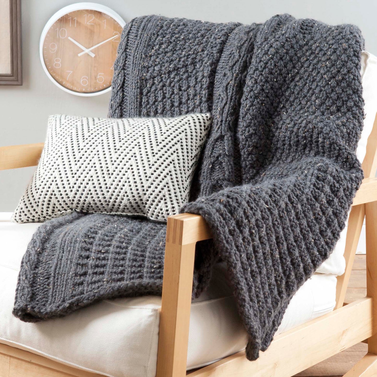 The Loom Knit Samplers blanket by Leisure Arts, featuring unique stitch patterns in gray, is draped over a light wooden armchair with a cream cushion. A white pillow with a black chevron pattern and a round wall clock complete the scene.