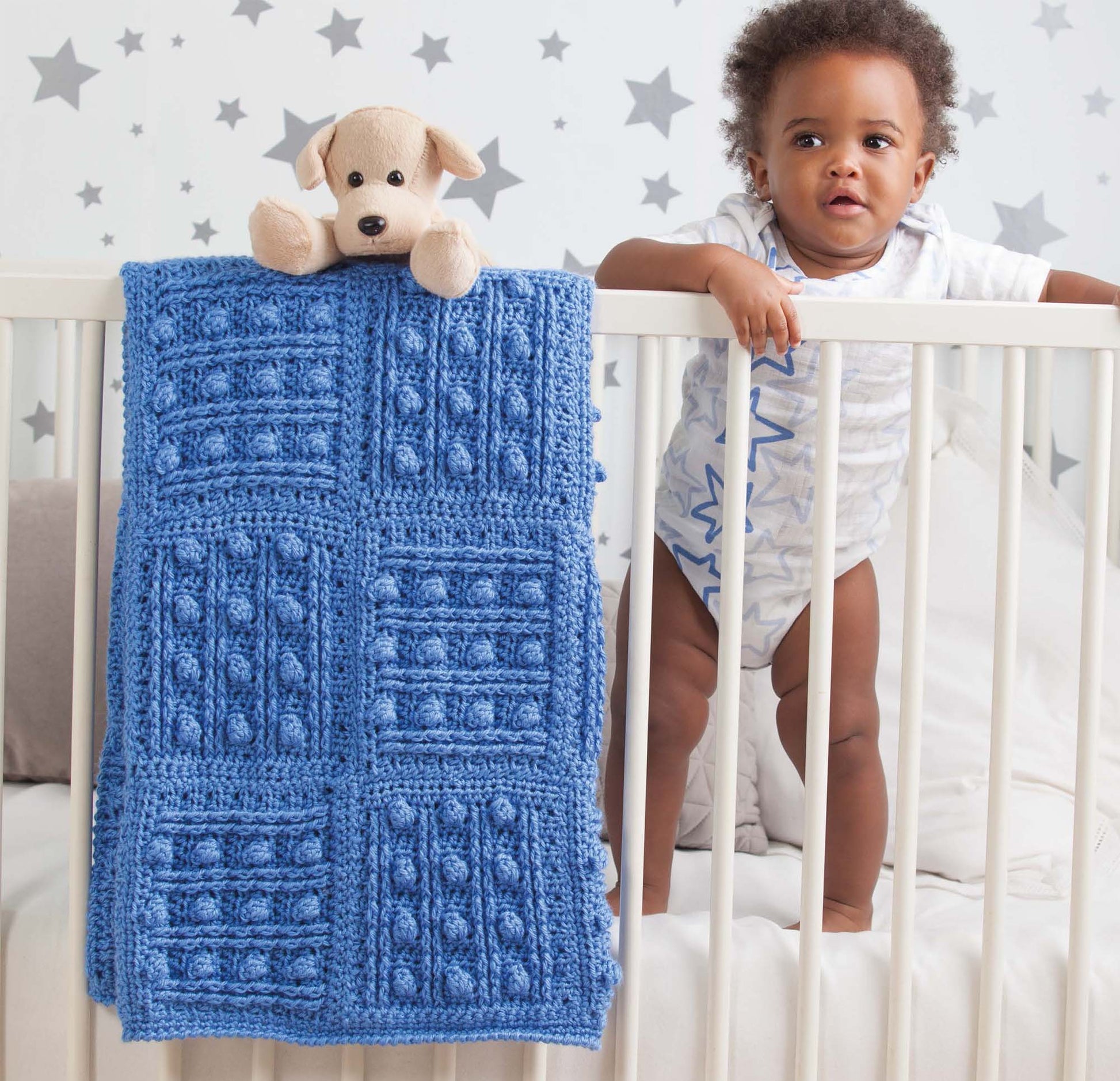 A baby stands in a white crib with a Leisure Arts Mix & Match Baby Sampler crochet blanket draped over the rail. A beige stuffed dog sits nearby, and the nursery wall features gray and white stars.