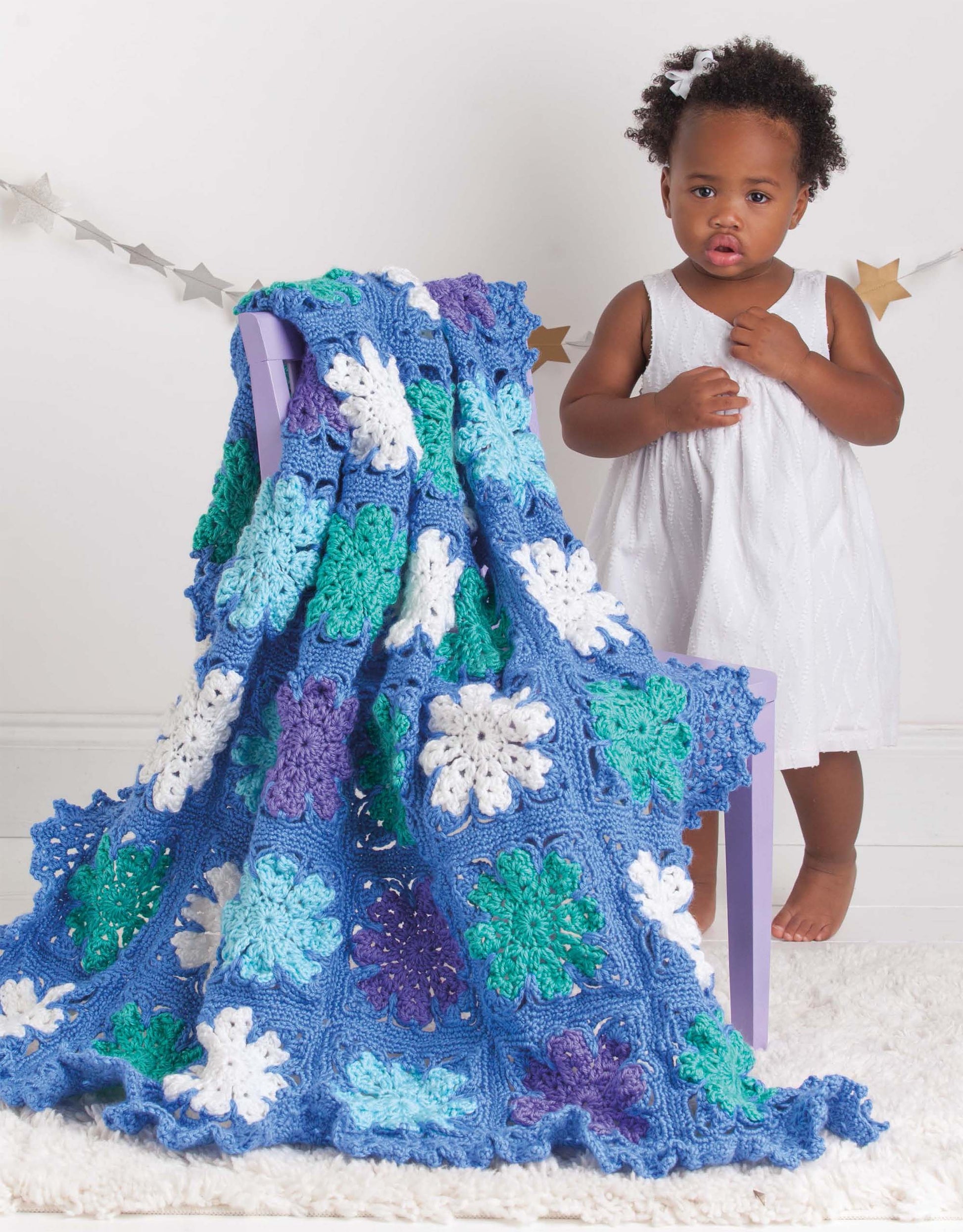 A young child in a white dress stands beside a purple chair draped with the Leisure Arts Mix & Match Baby Sampler, a colorful crocheted blanket featuring blue, green, white, and purple granny squares. A star garland decorates the simple background.