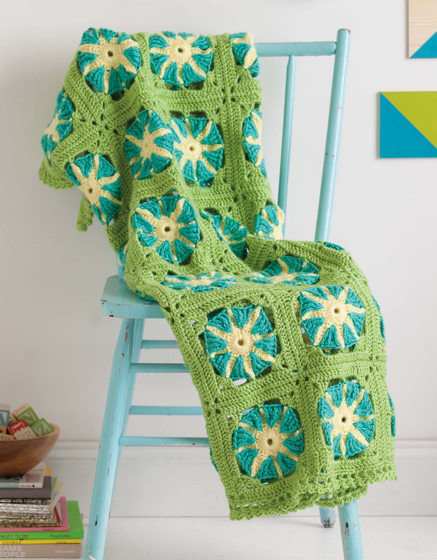 A Mix & Match Baby Sampler crocheted blanket by Leisure Arts with green, blue, and yellow granny squares drapes over a light blue chair next to books and a wooden bowl in a bright, modern room.