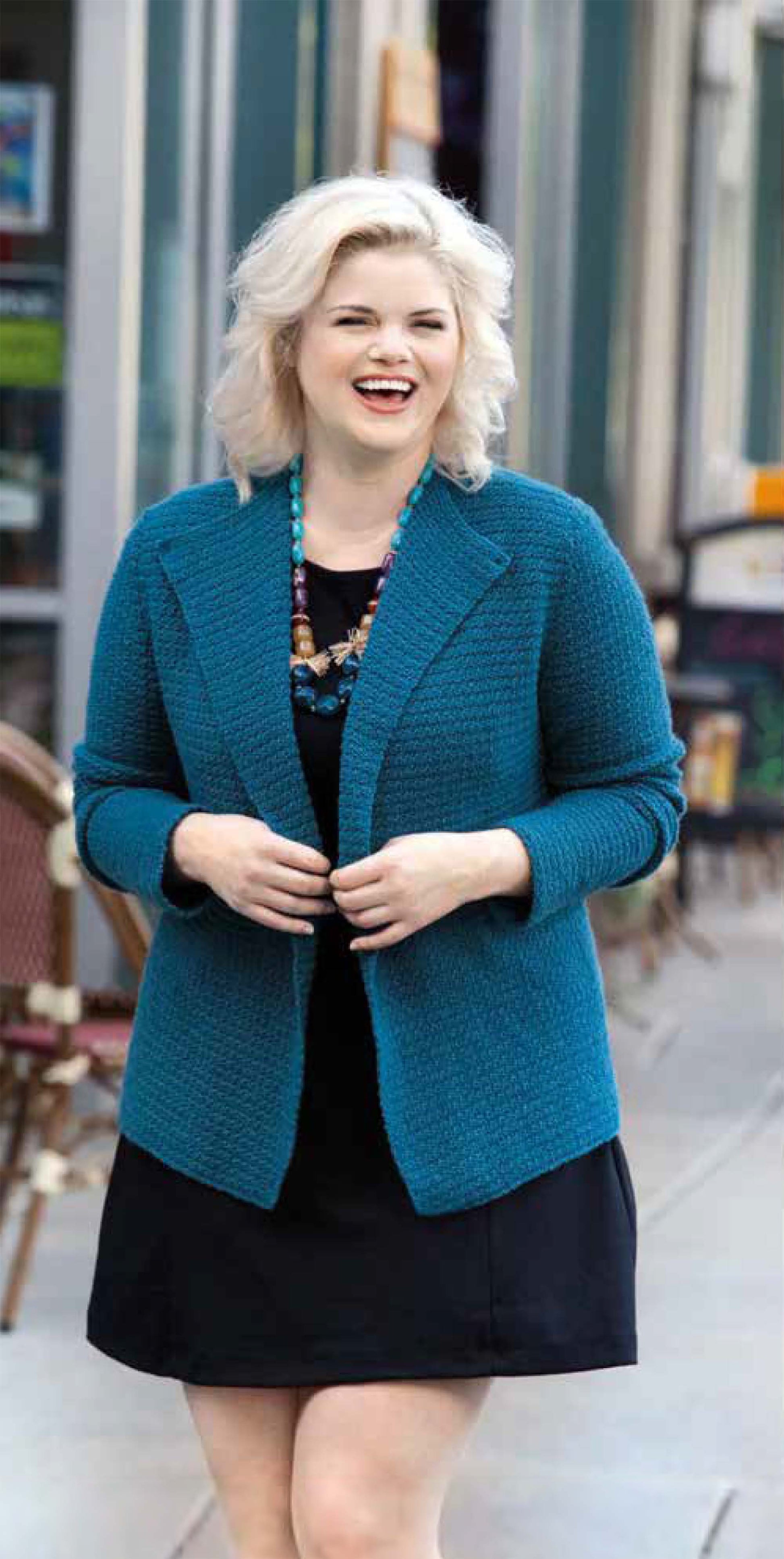 A smiling blonde woman wears a teal knit blazer and black dress made from Leisure Arts' Plus-Size Garments with Style, paired with a blue beaded necklace, standing on a city sidewalk near outdoor cafe seating.