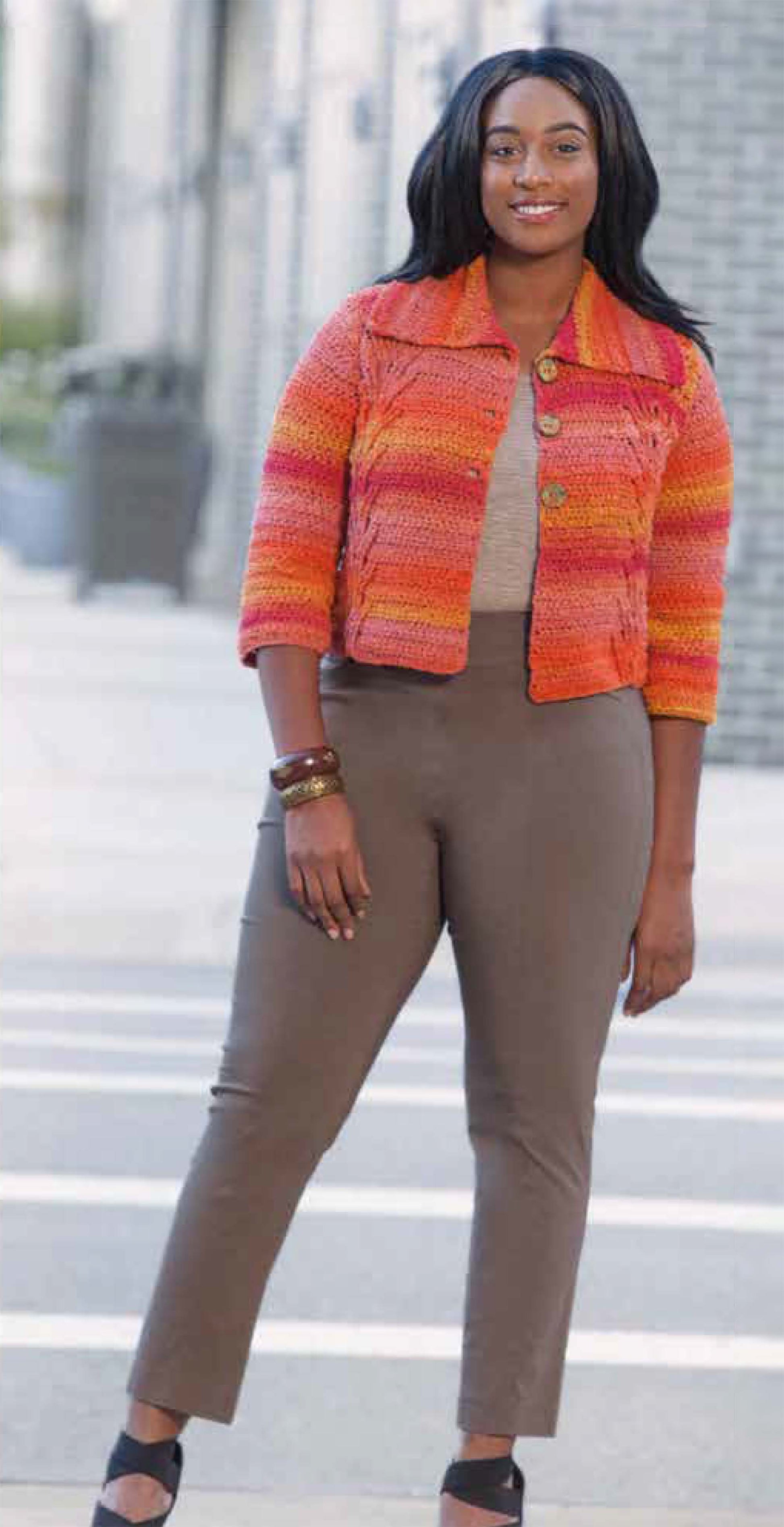 A woman stands outdoors smiling, wearing a vibrant orange and pink jacket from Leisure Arts' Plus-Size Garments with Style collection over a beige top and brown pants, radiating cheerful confidence.