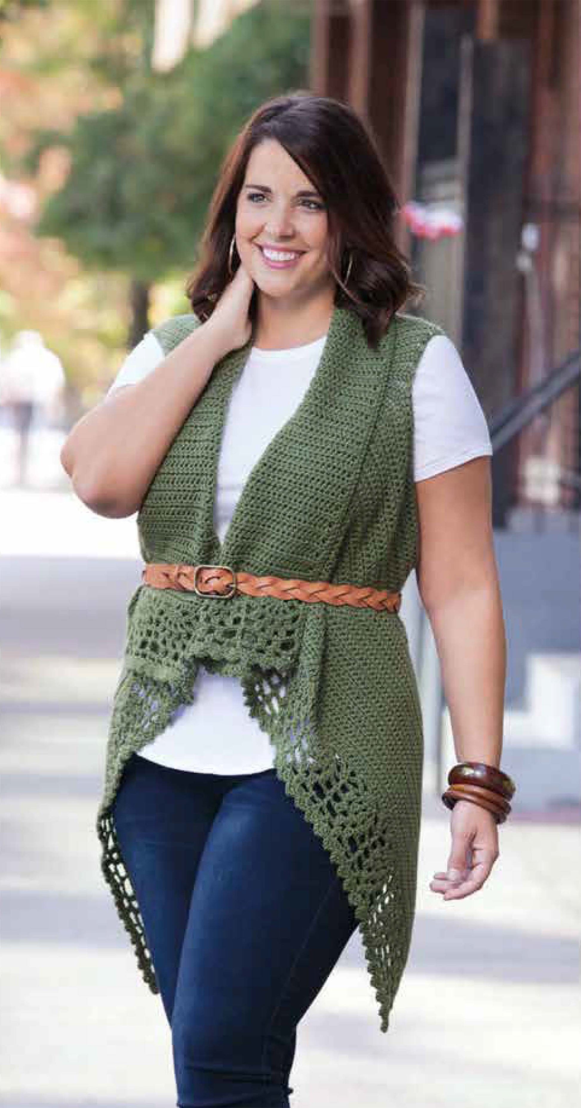 A woman in a white shirt, blue jeans, and a green vest from the Plus-Size Garments with Style collection by Leisure Arts smiles while walking outdoors on a sunny day, accessorized with a brown braided belt and bracelets.