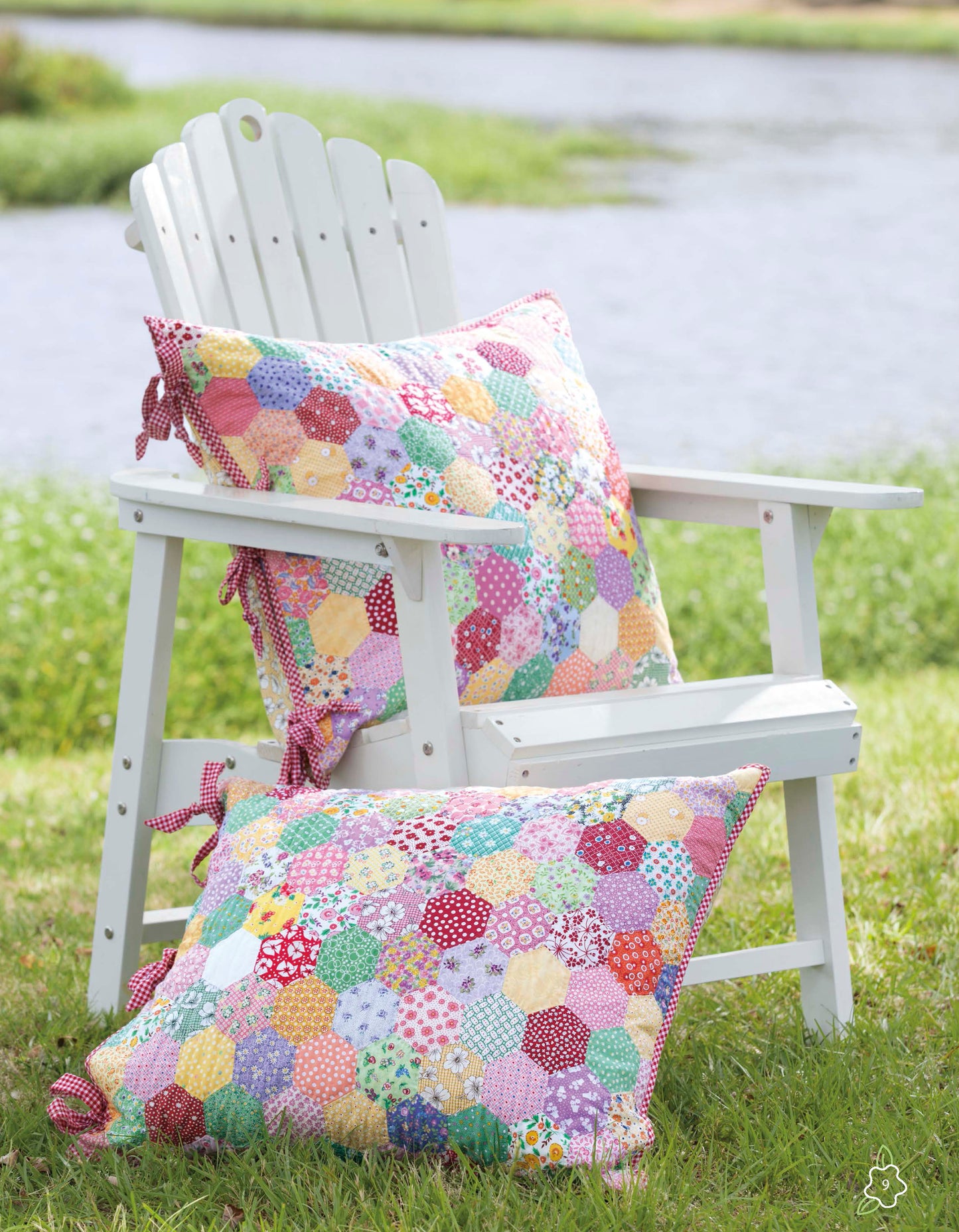 A white Adirondack chair featuring two vibrant patchwork pillows from the Salt Water Taffy Collection by Leisure Arts, inspired by English paper piecing and heirloom quilts, sits on grass beside a pond with water and greenery in the background.