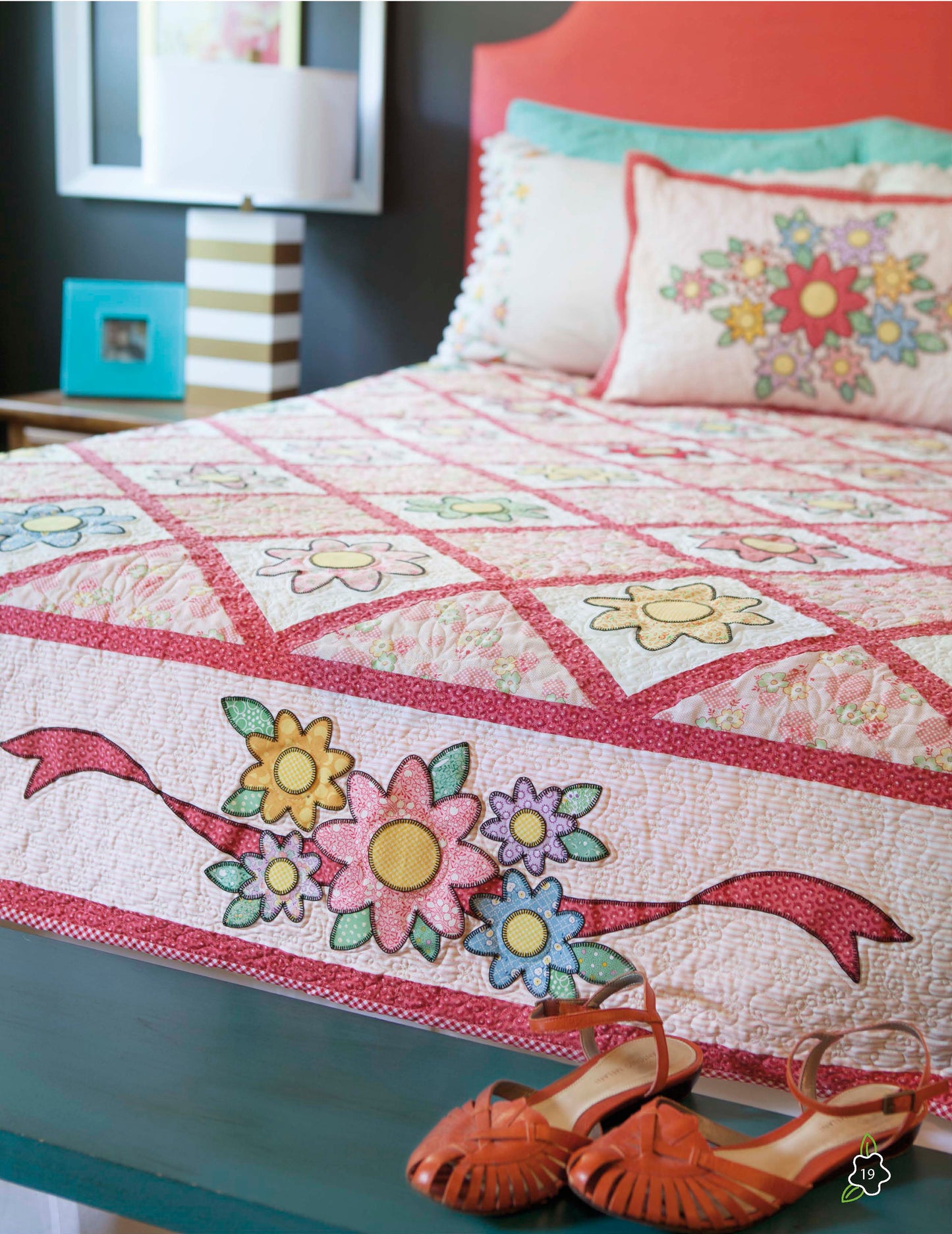 A bed dressed in the vibrant Salt Water Taffy Collection quilt by Leisure Arts, featuring English paper piecing and delicate hand embroidery, stands out against a coral headboard. Orange shoes are placed atop a dark green bench at the foot of the bed, while a striped lamp and photo frame decorate the nearby bedside table.